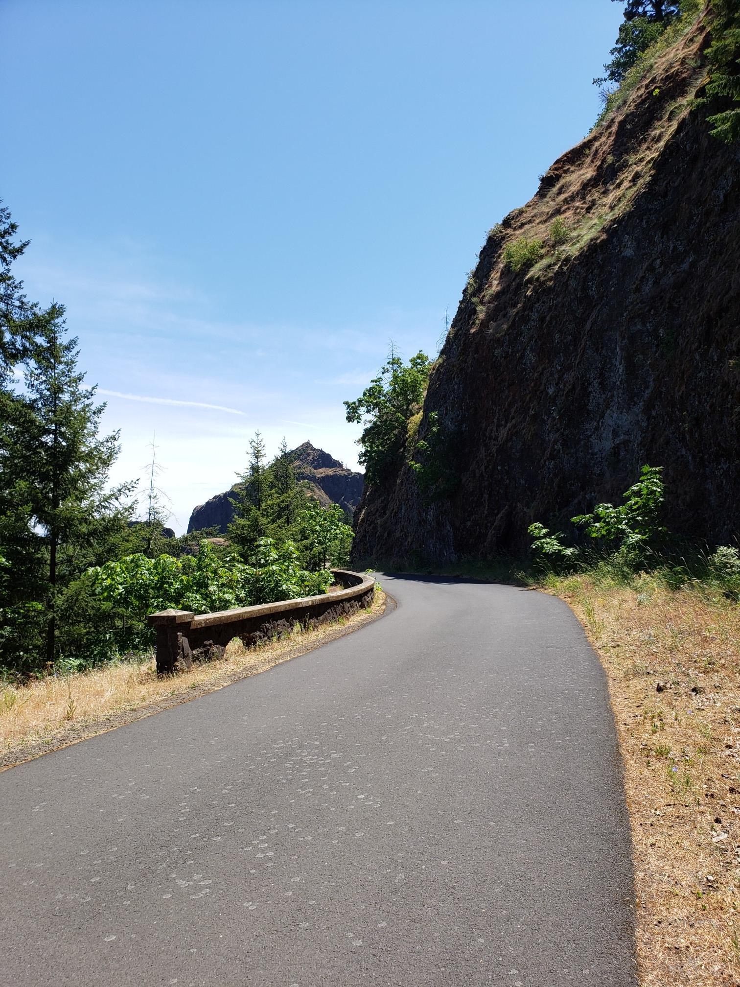 Paved road curves beside a steep rock wall on a sunny day.