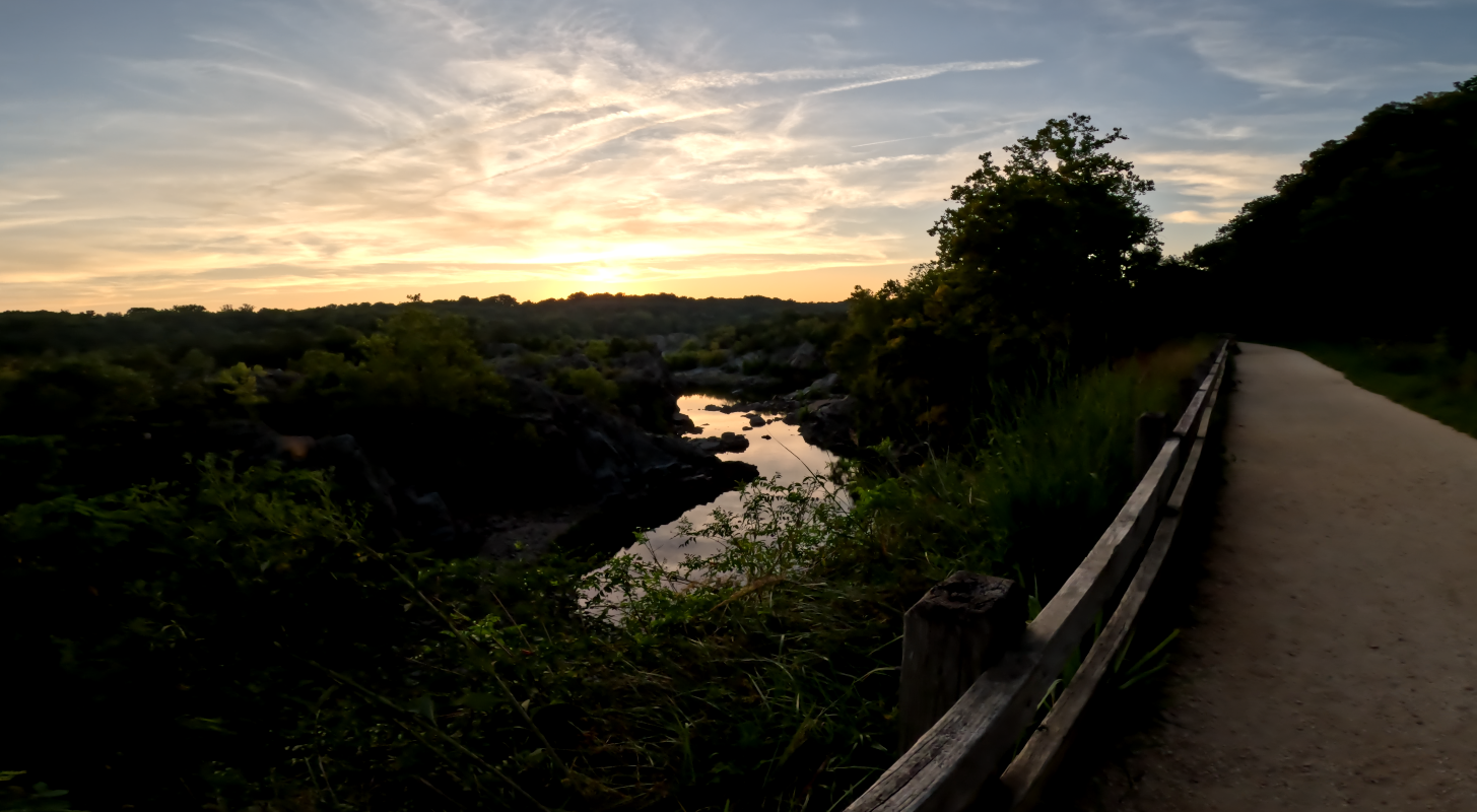 A dirt path and wooden fence beside a rocky river at sunset.