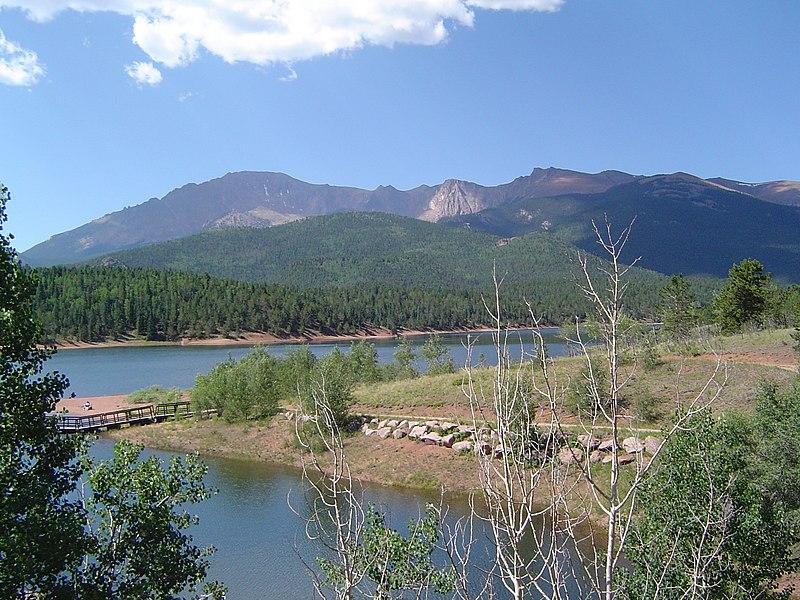 A sunny lake with a dock, surrounded by pine forests and distant mountains.