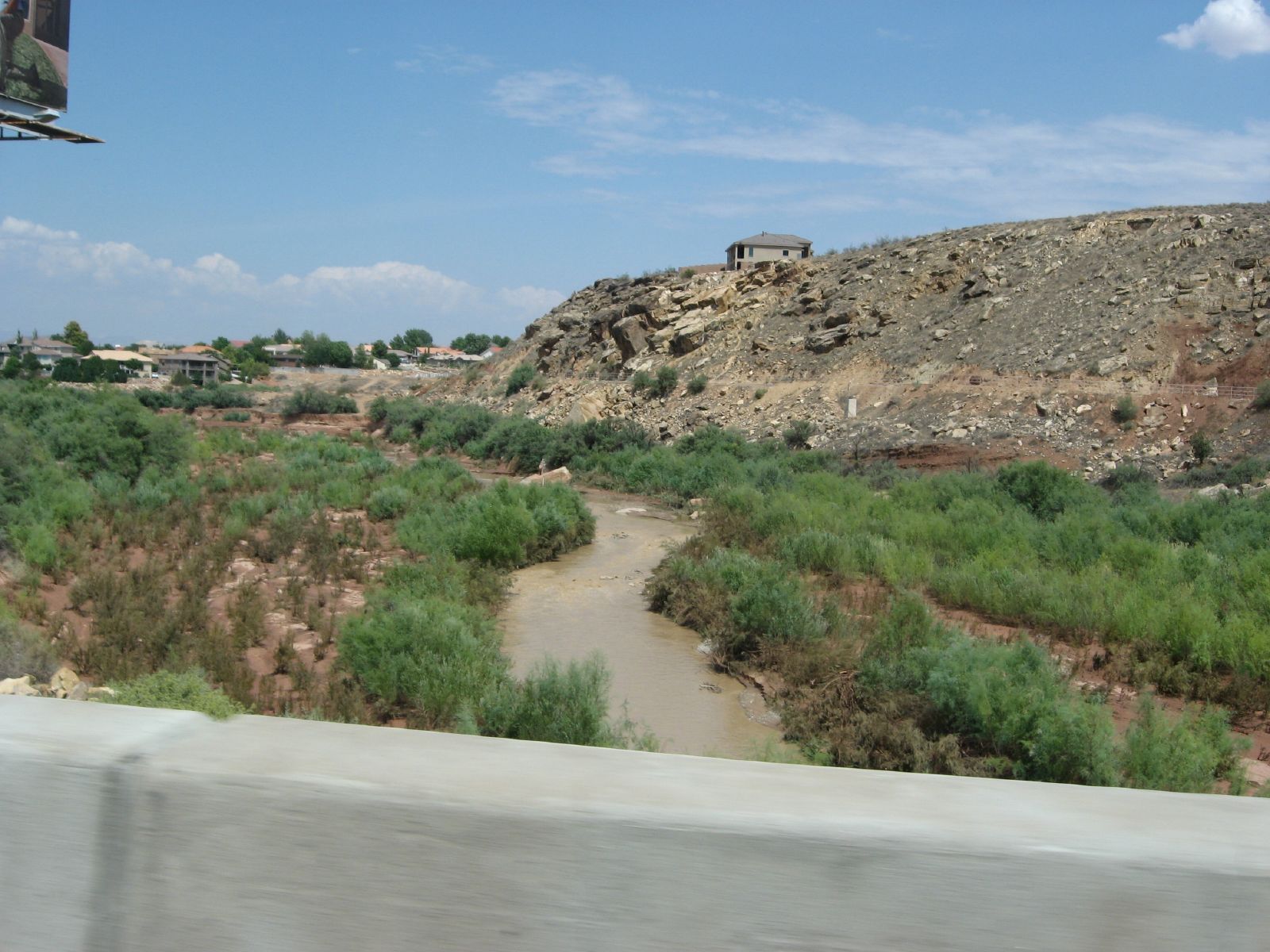 A dry riverbed with green bushes, rocky hills, and distant houses under a bright sky. - Bloomington Park – Views of Virgin River loop from Practice Sand Trap