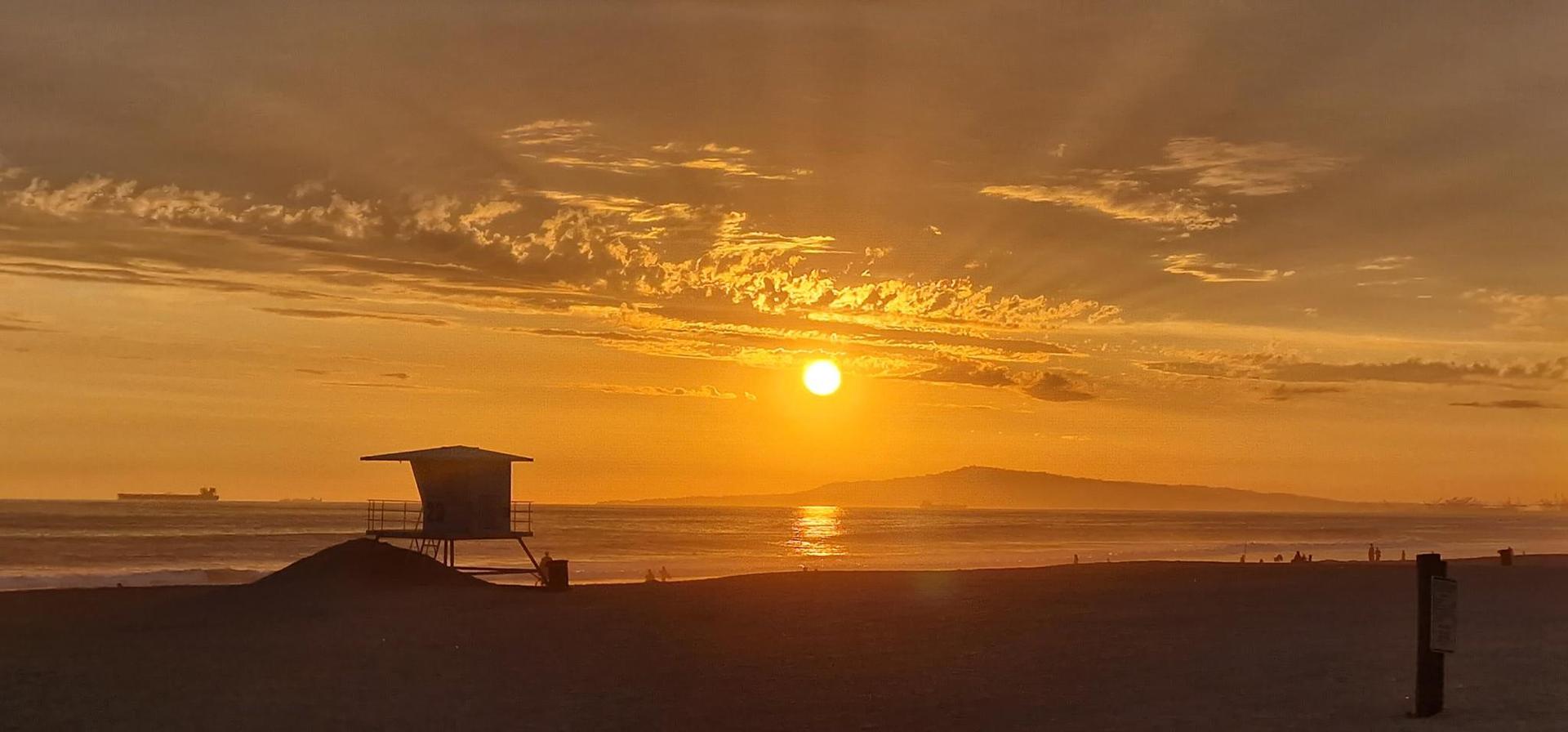 Sunset over the ocean with a lifeguard tower silhouette and distant mountain. Golden light.
