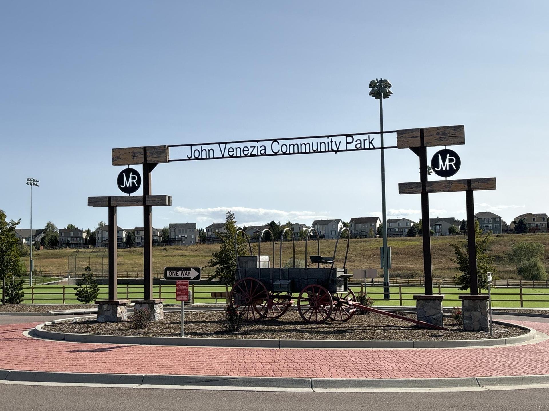 An entrance sign over a decorative wagon, with houses and fields in the background.