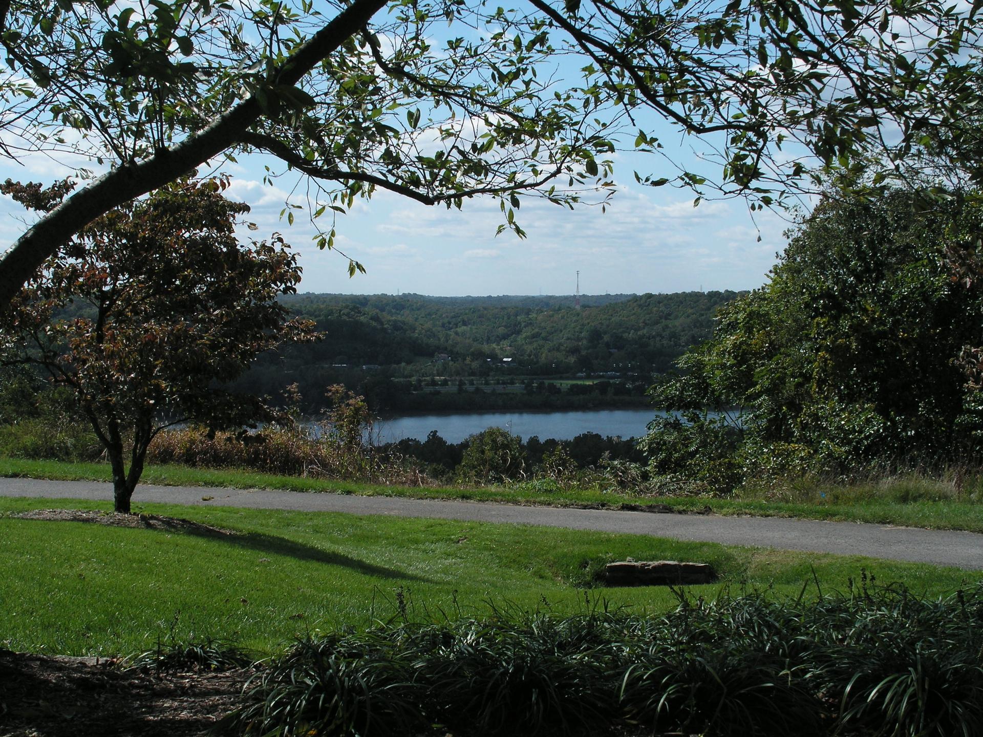 A sunny overlook showing a lake, forested hills, and a grassy path.