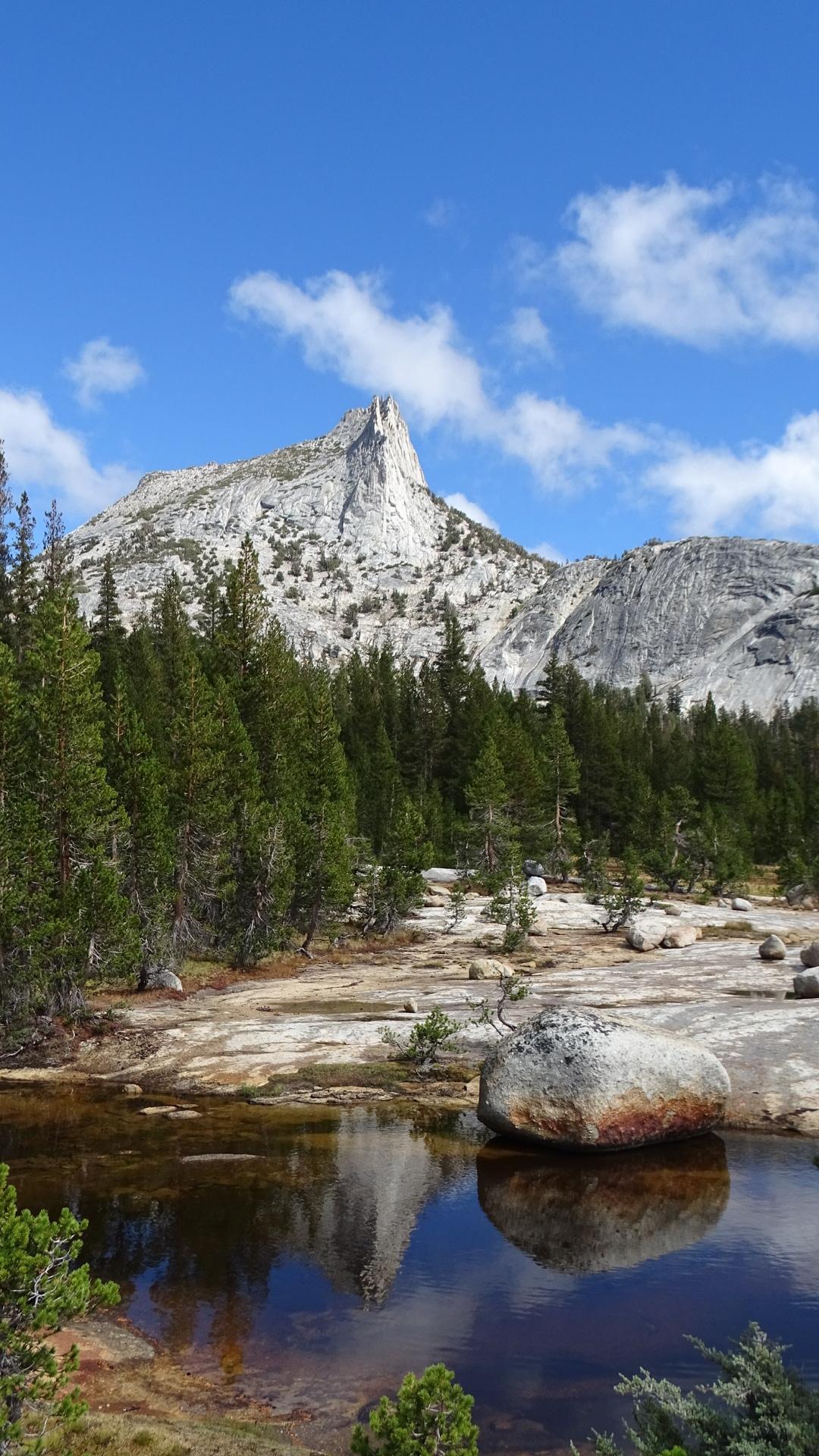 Jagged mountain peak reflected in calm water, surrounded by pine trees under blue sky.
