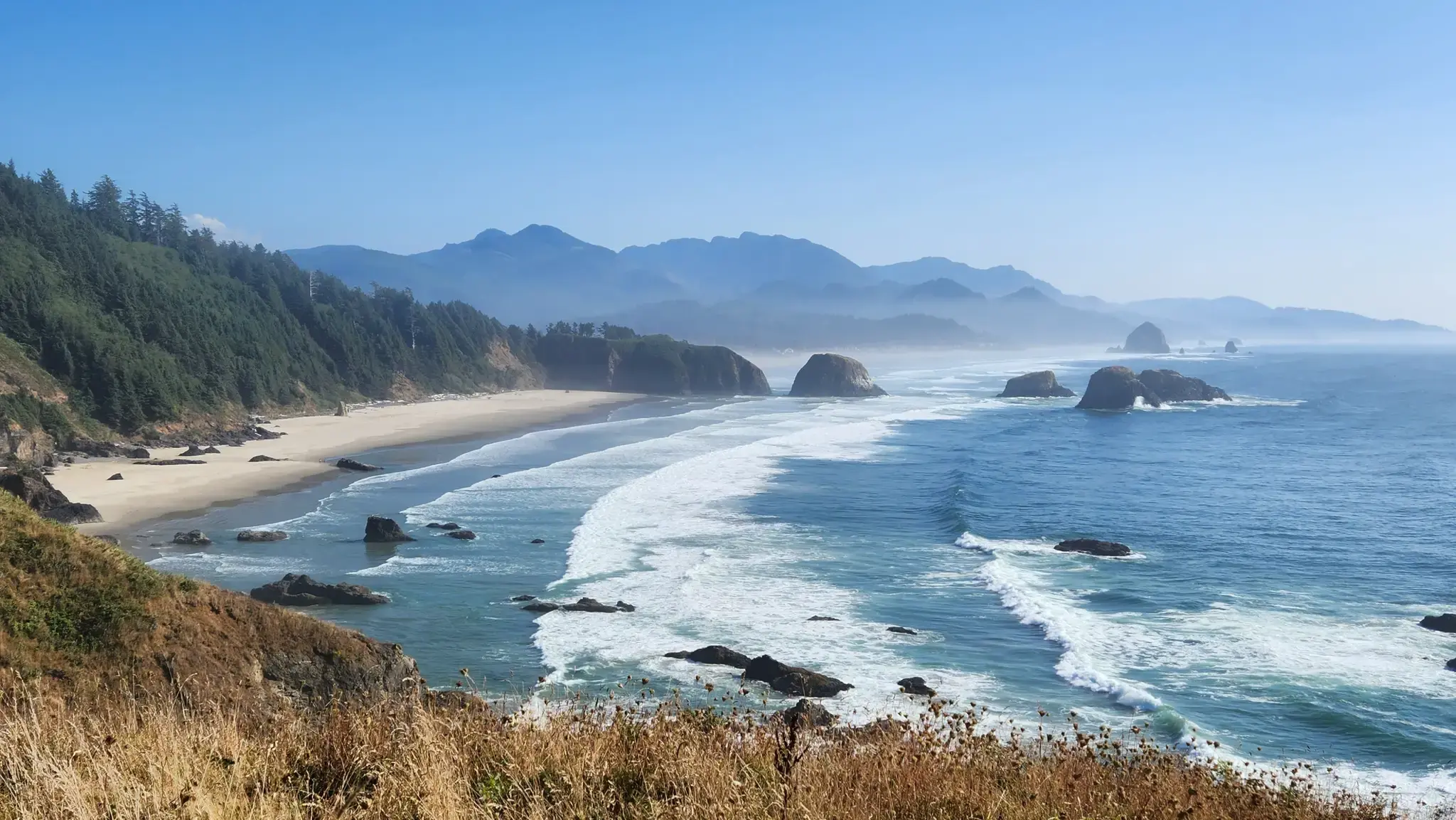 Sunny panoramic view of a wide beach, ocean, and distant misty mountains.