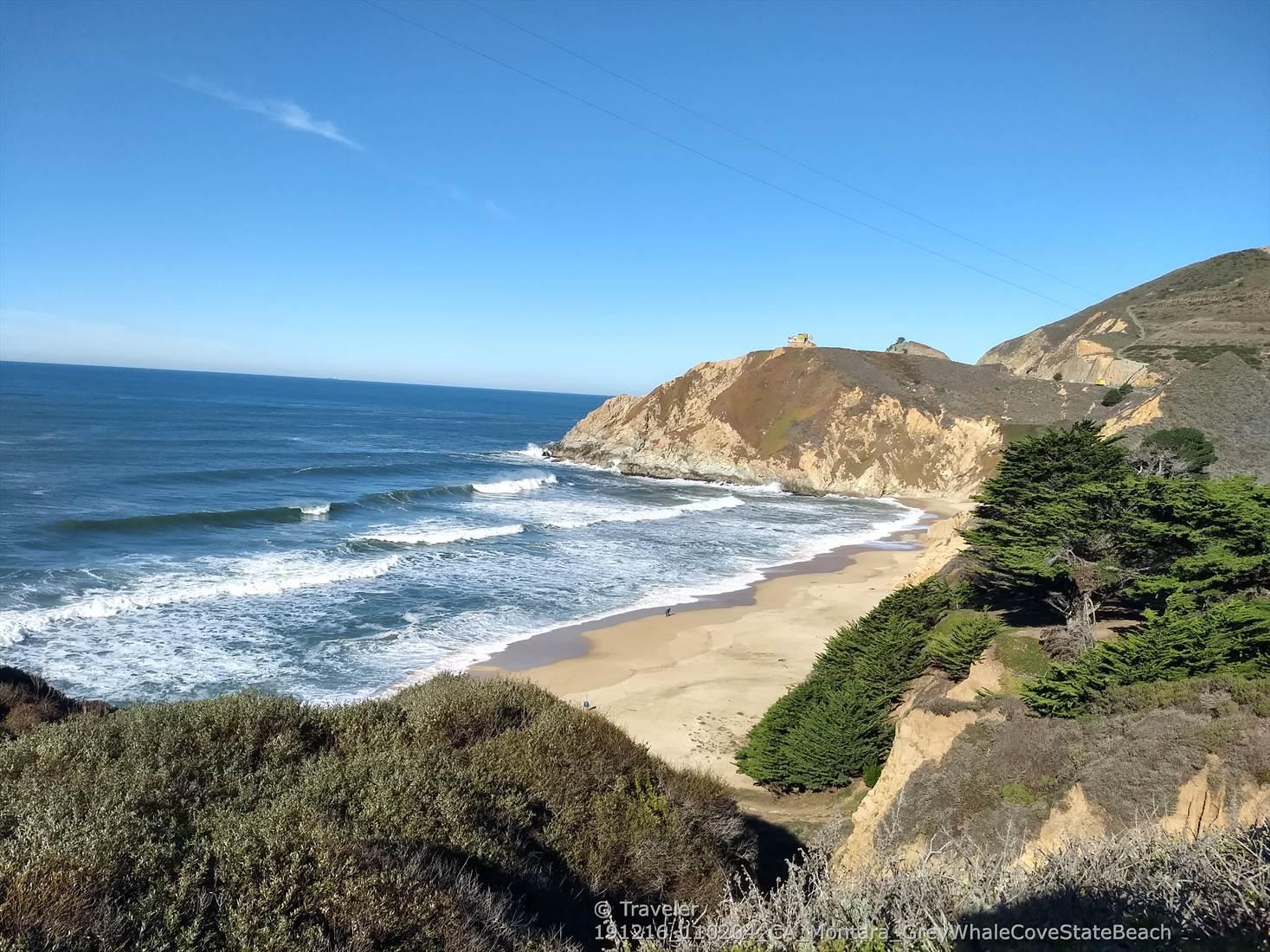 Sunny beach with crashing waves, rocky cliffs, and green trees.
