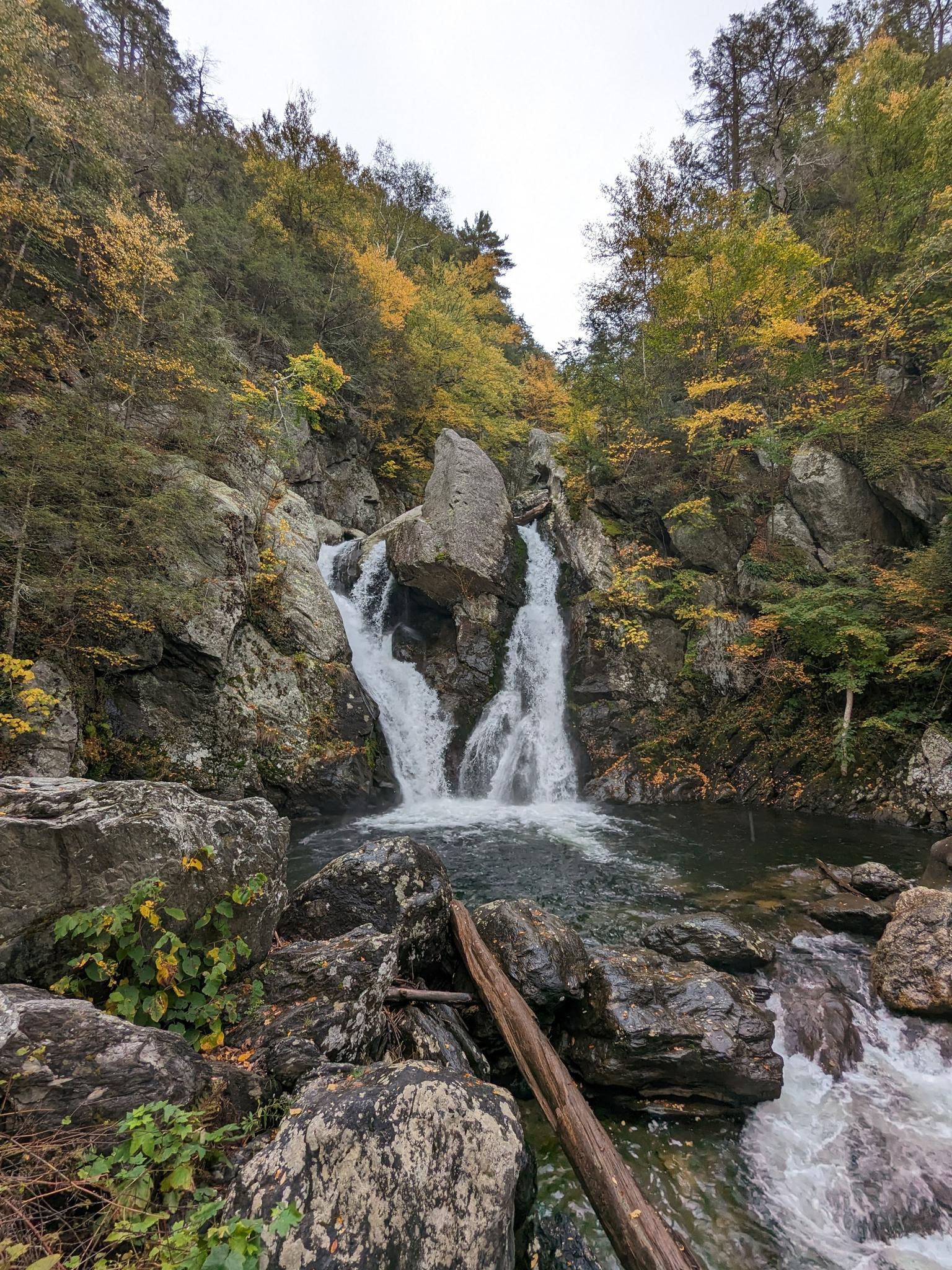 Double waterfall cascades into a rocky pool surrounded by autumn trees.