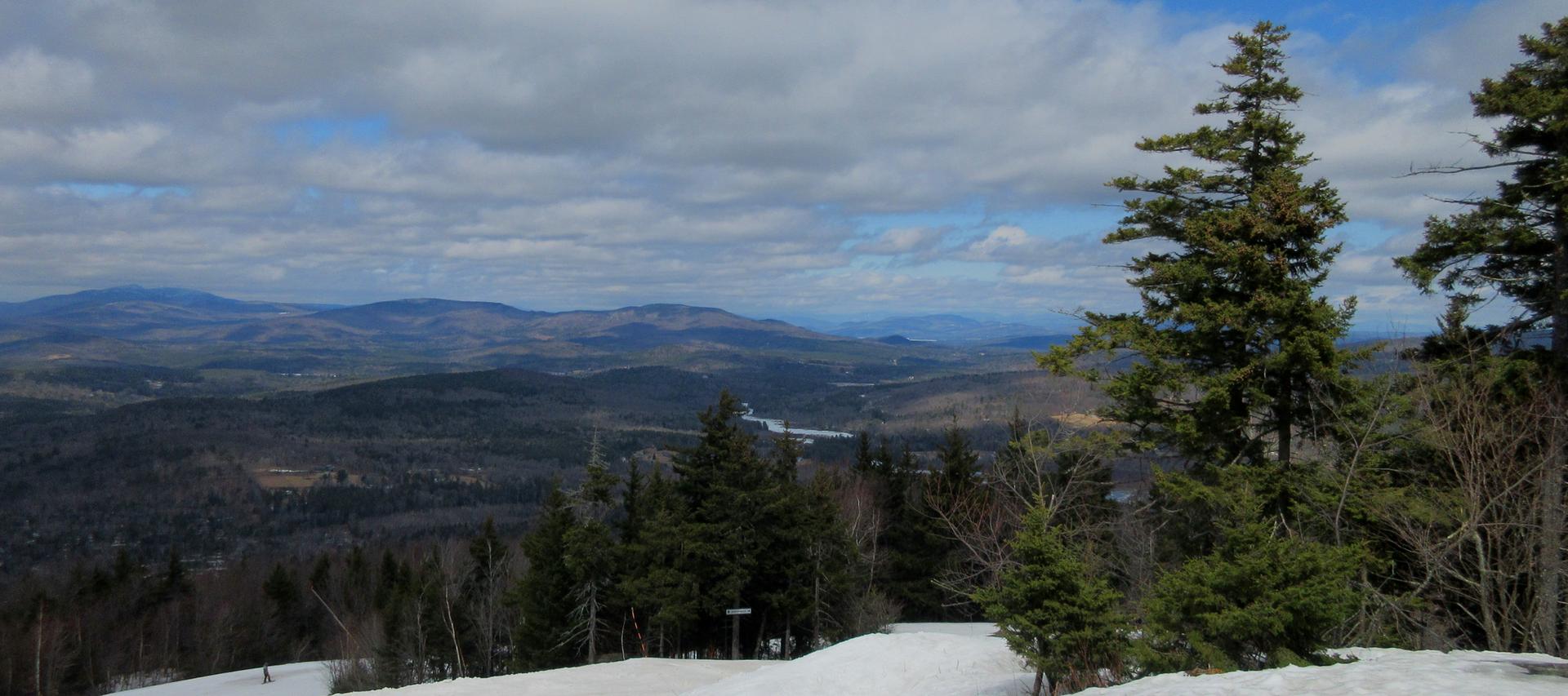 Panoramic mountain view with snow, evergreen trees, and distant hills under a cloudy sky.