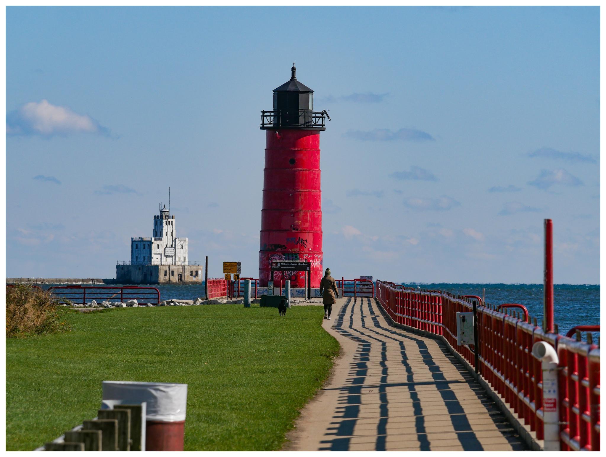 A red lighthouse on a pier with people walking, under a clear sky.
