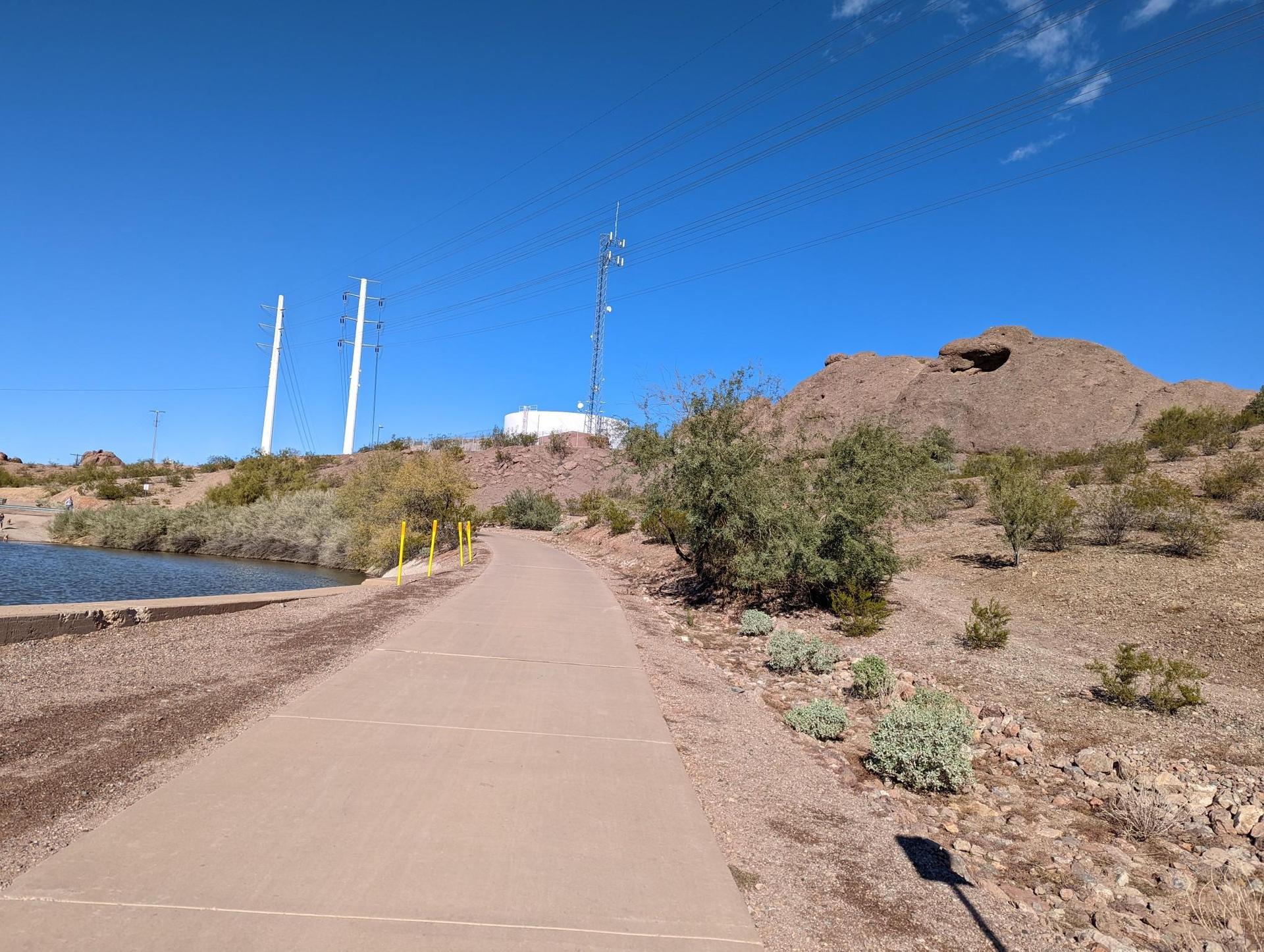 Paved path alongside water, desert hills, utility poles, bright sunny day.