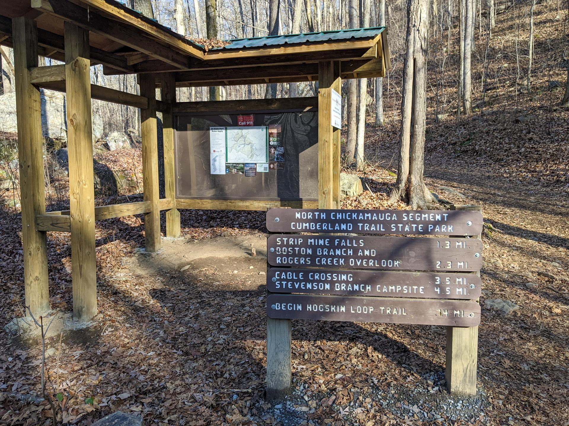 A wooden information kiosk and trail sign stand in a sunny, bare forest.