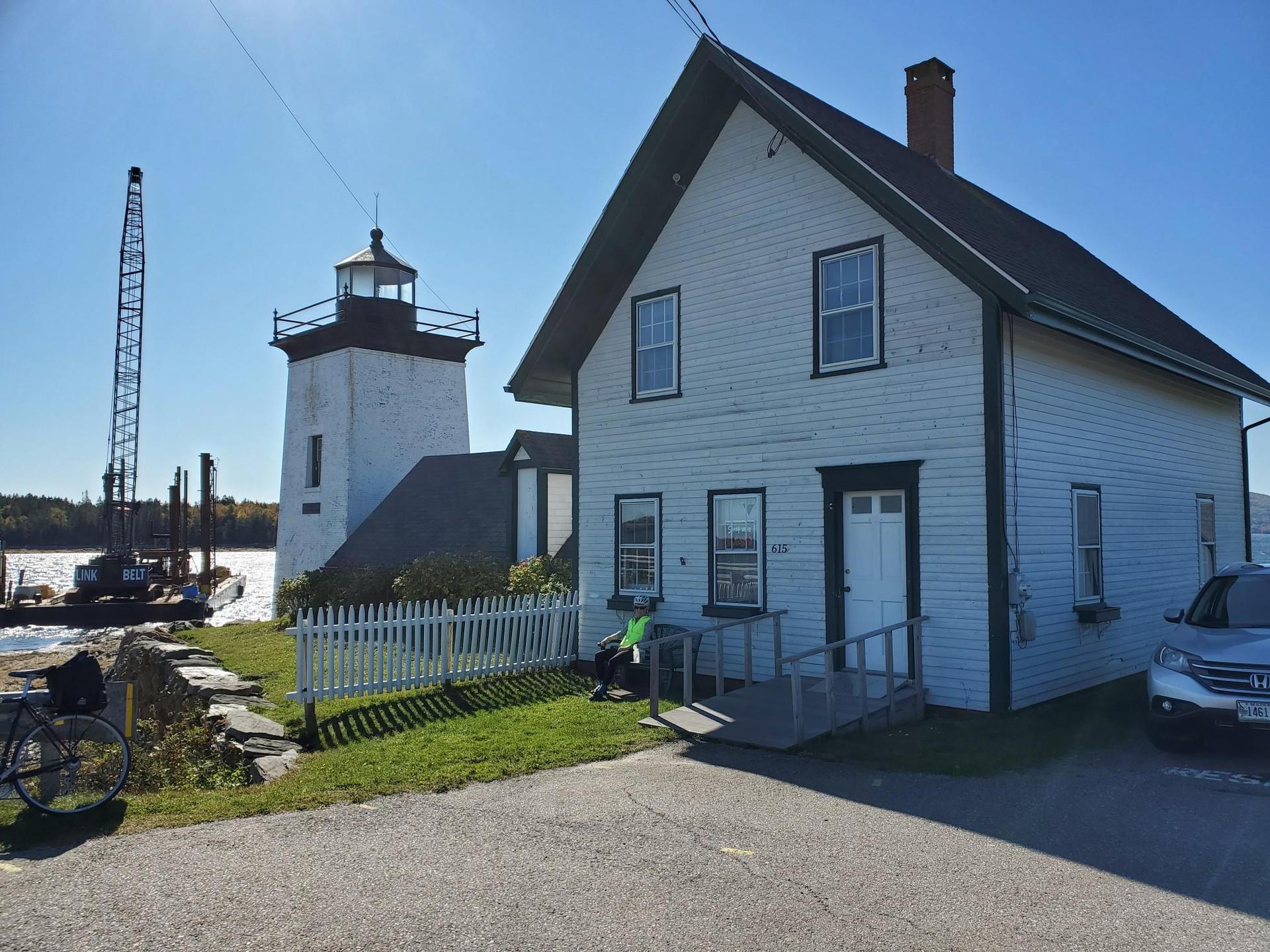 White house and lighthouse by a sunny waterfront with a crane and boat.