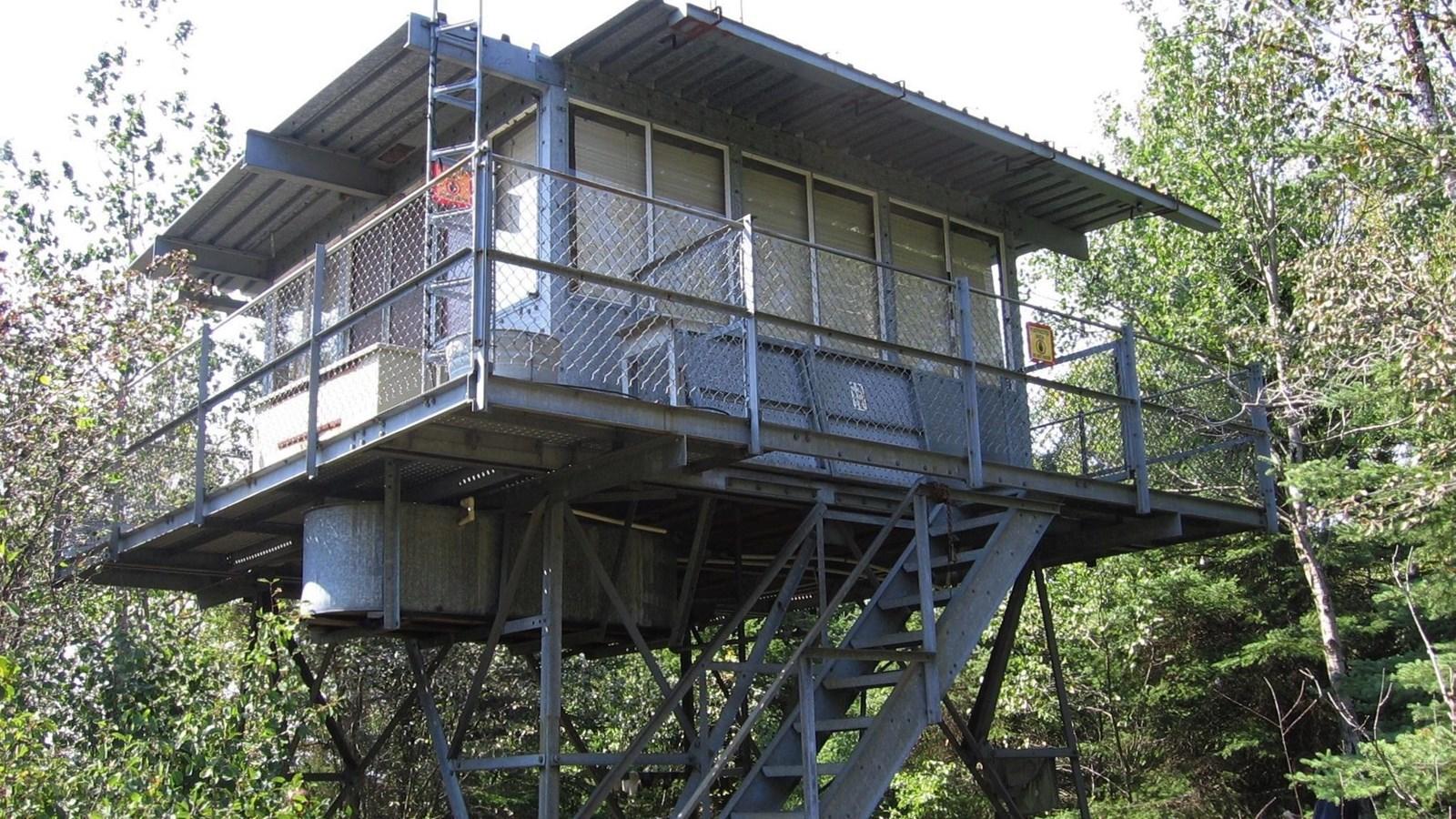 A metal fire lookout cabin stands high among green trees on a bright day.