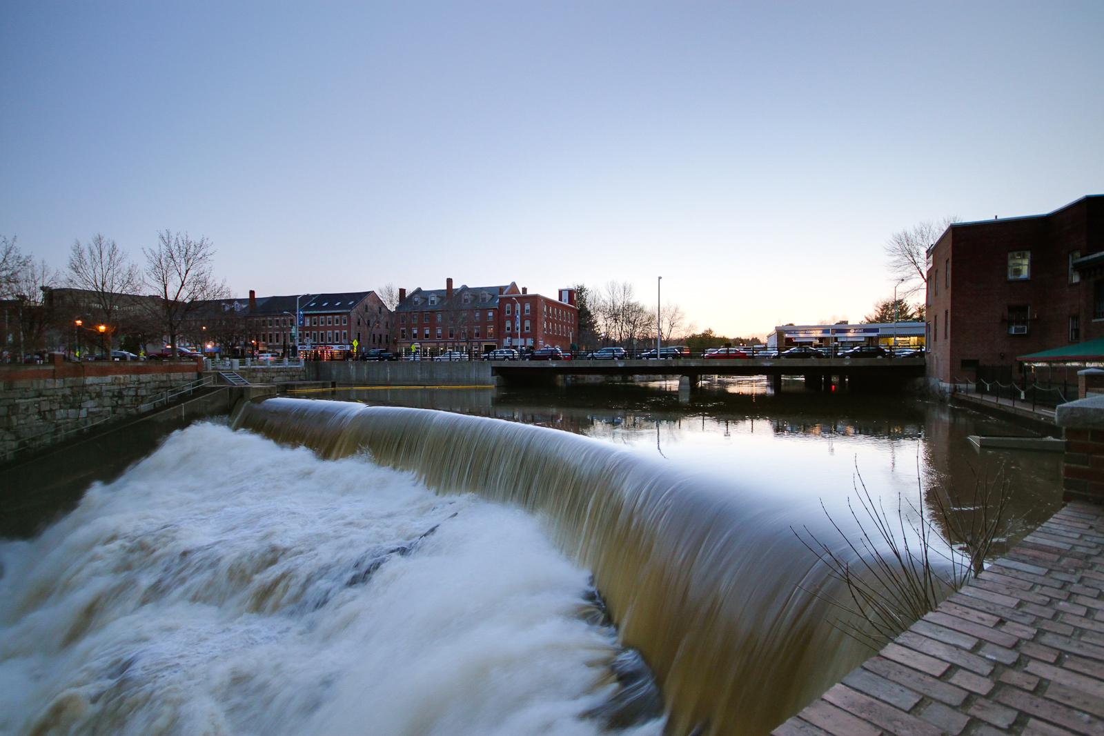 Waterfall in a river with urban buildings and a bridge at dusk.