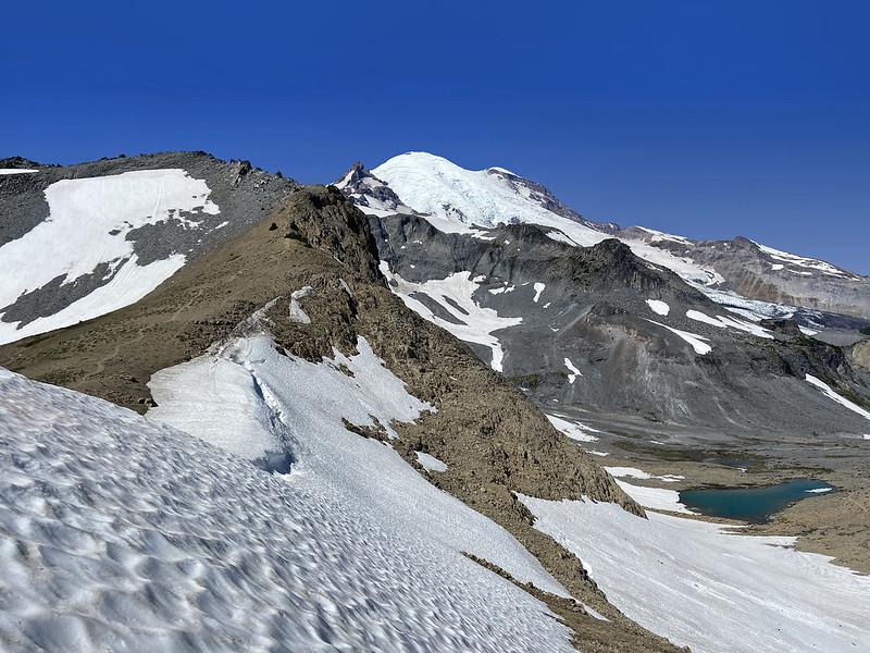 Snowy mountain peak, rocky ridge, and small lake under clear sky.