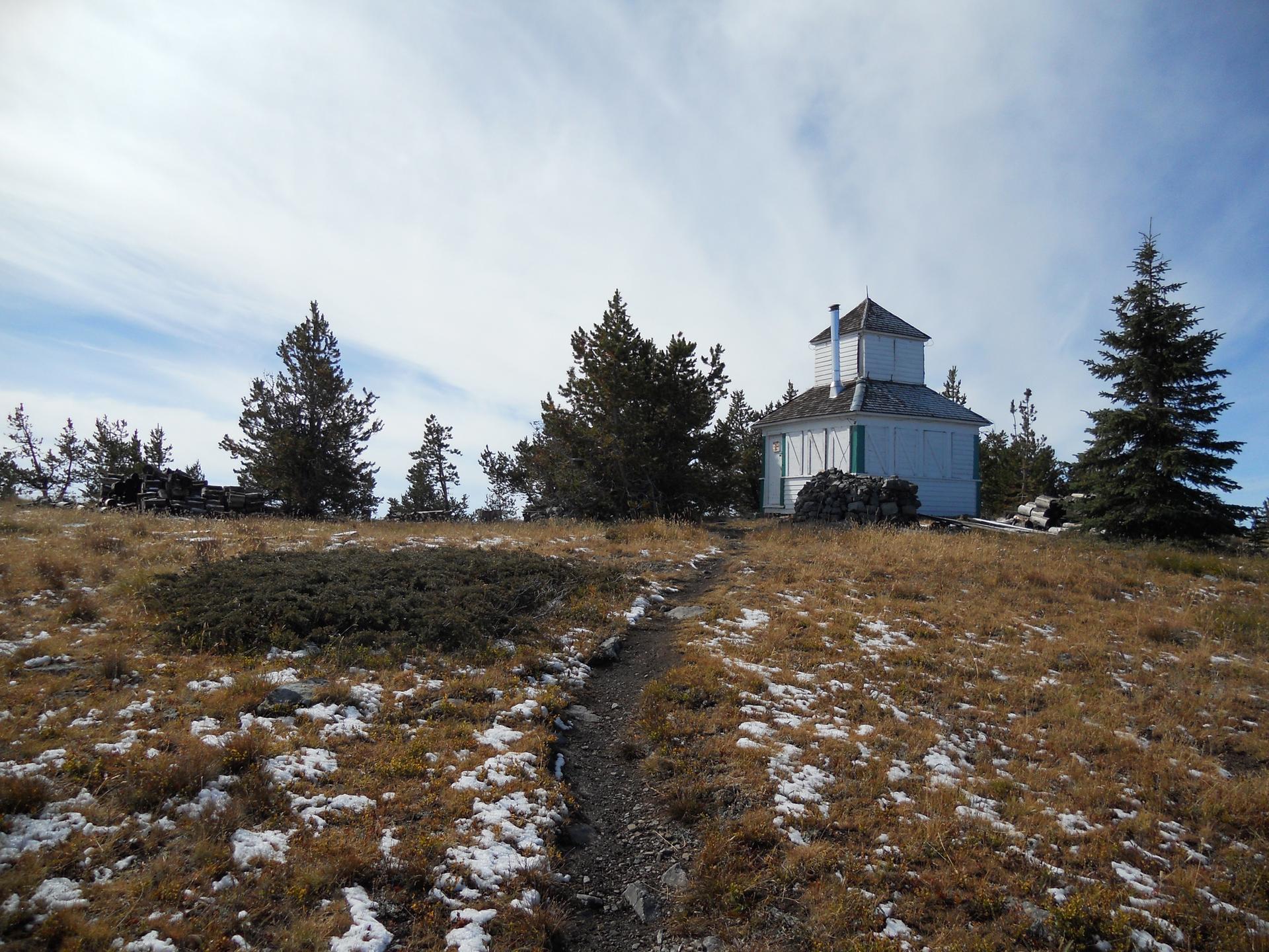 Dirt path leading to a white building on a snowy, grassy hill with pine trees.