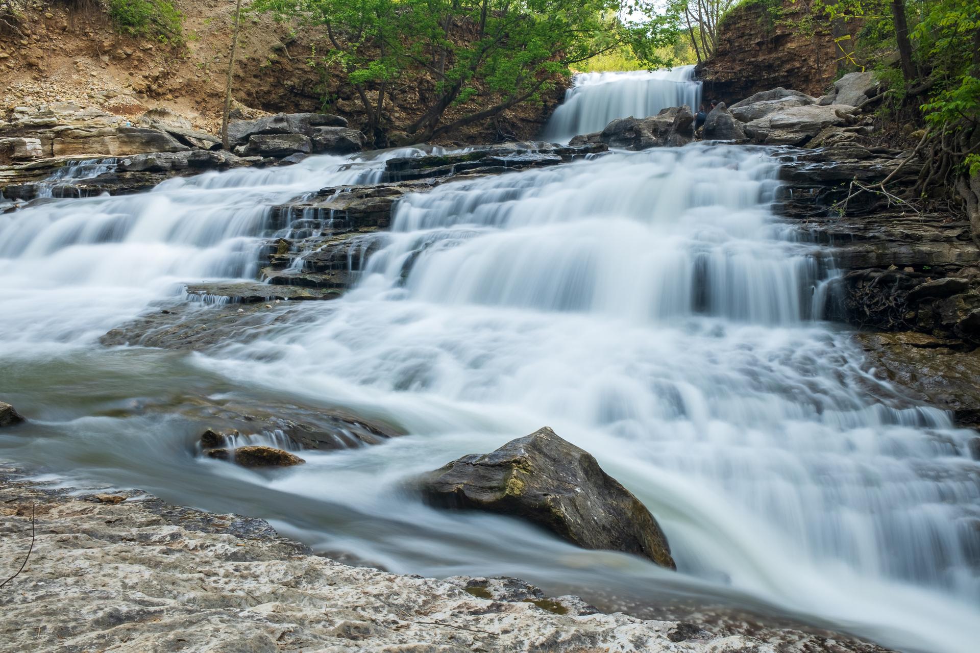 Long exposure waterfall cascades over rocky ledges with green trees above.