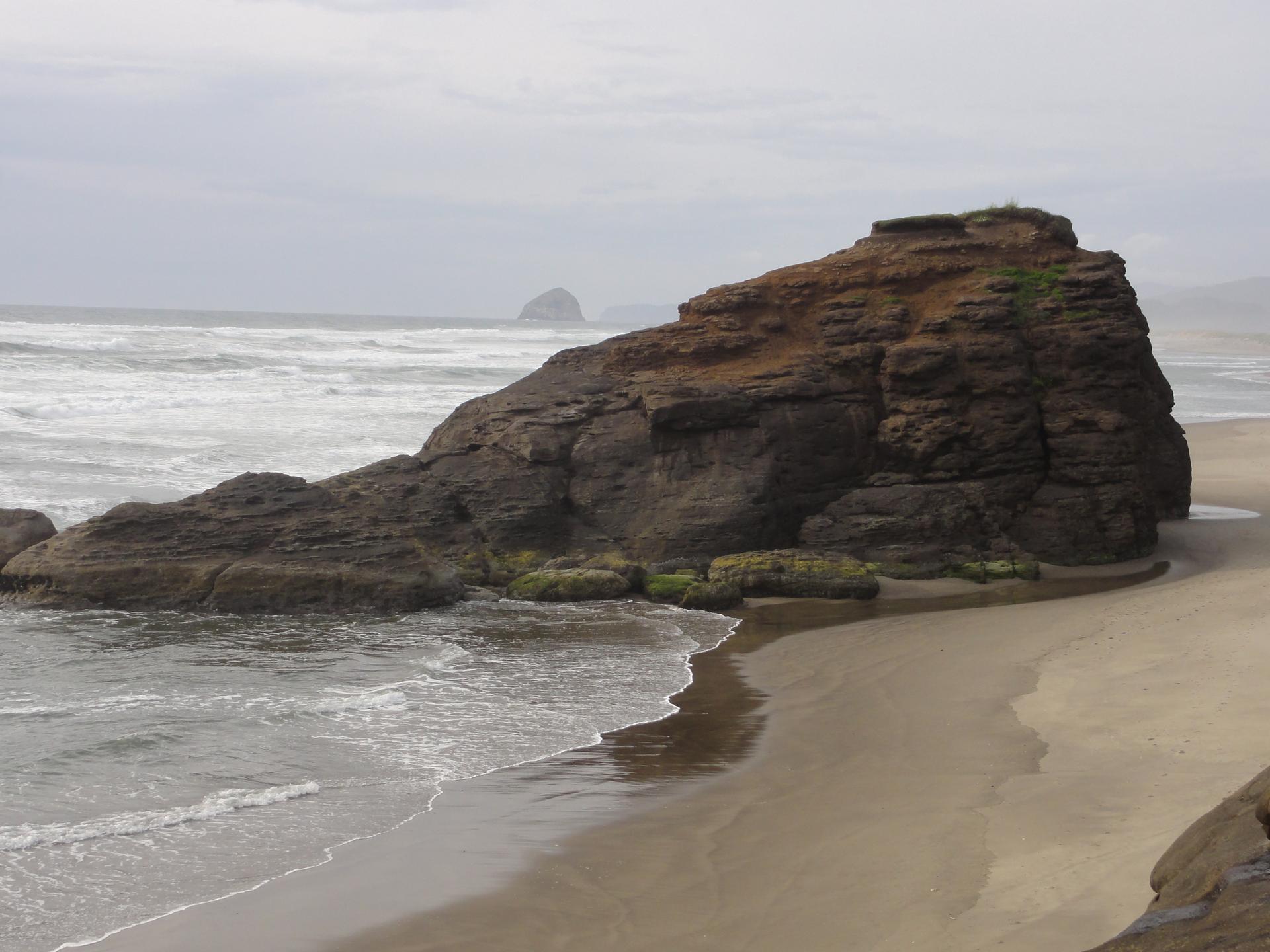 Large dark rock formation on a sandy beach with ocean waves under cloudy sky.