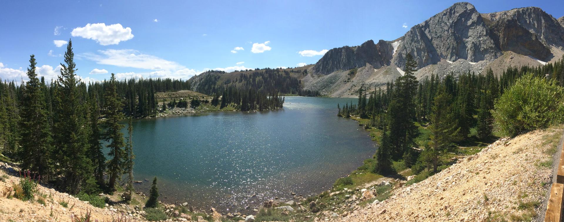 Panoramic view of a clear mountain lake surrounded by pine trees and rocky peaks.