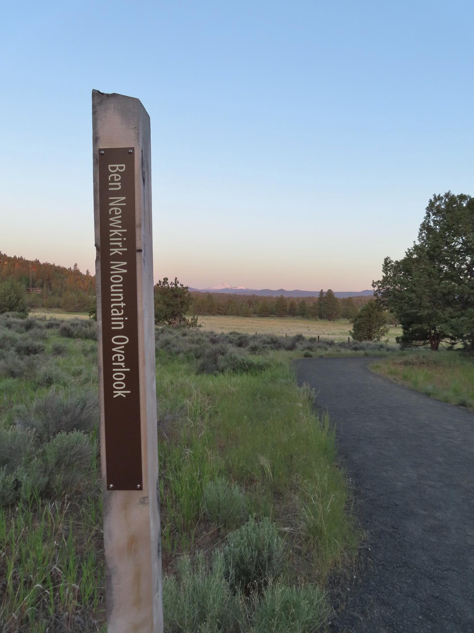 A signpost with text next to a dirt path overlooking a field at dawn.
