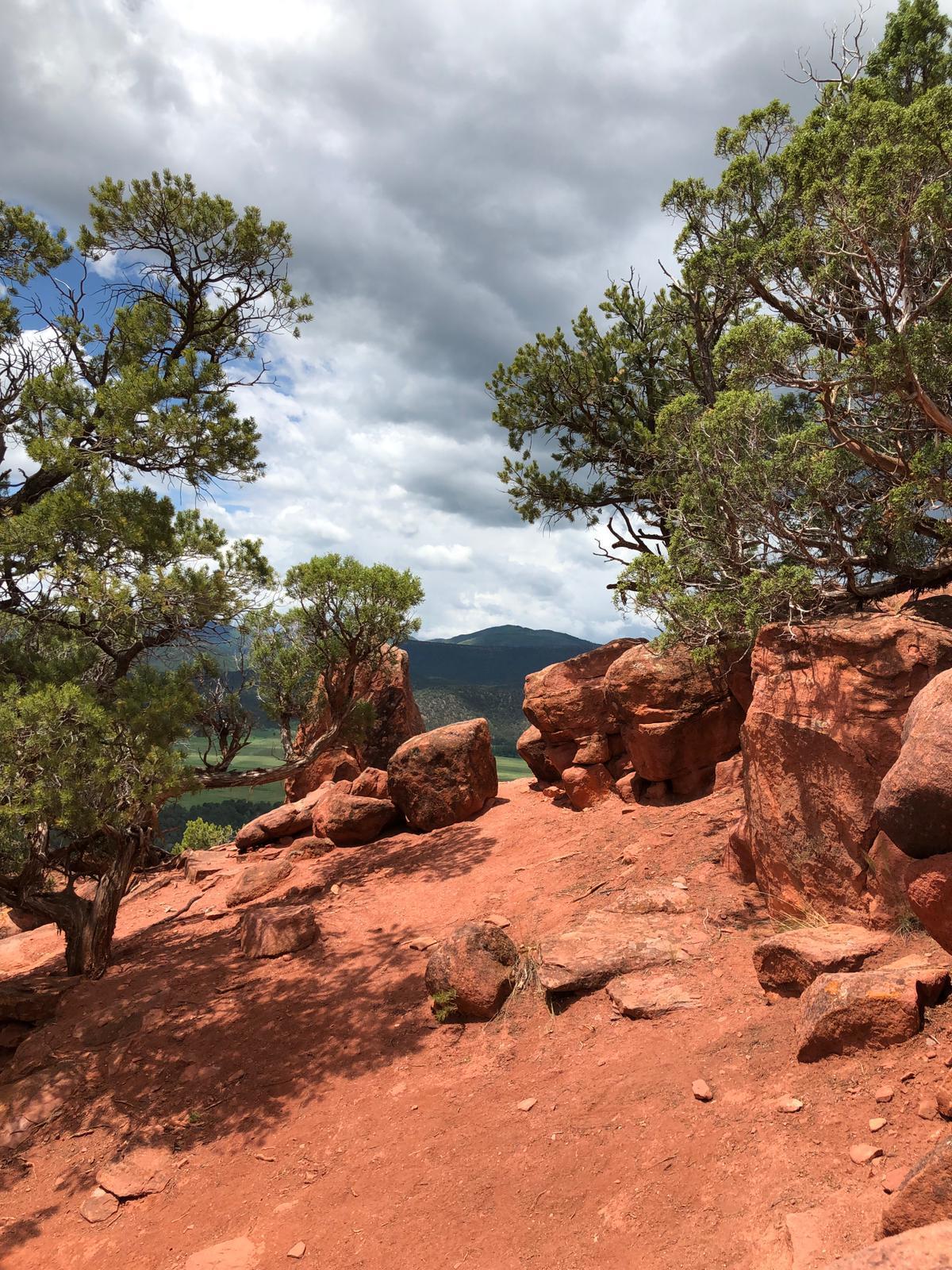 Red rock formations and trees on a hill, with green hills beyond.
