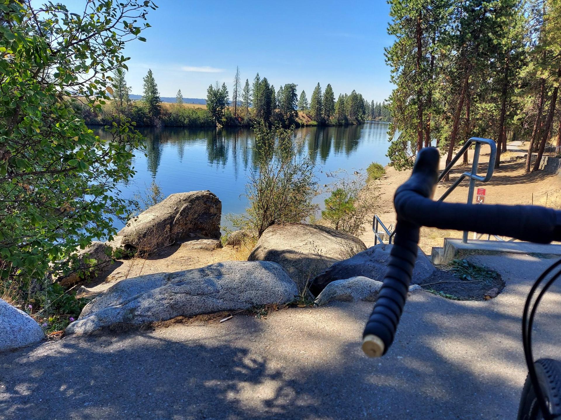 Bicycle handlebars overlooking a calm river, large rocks, and green trees.