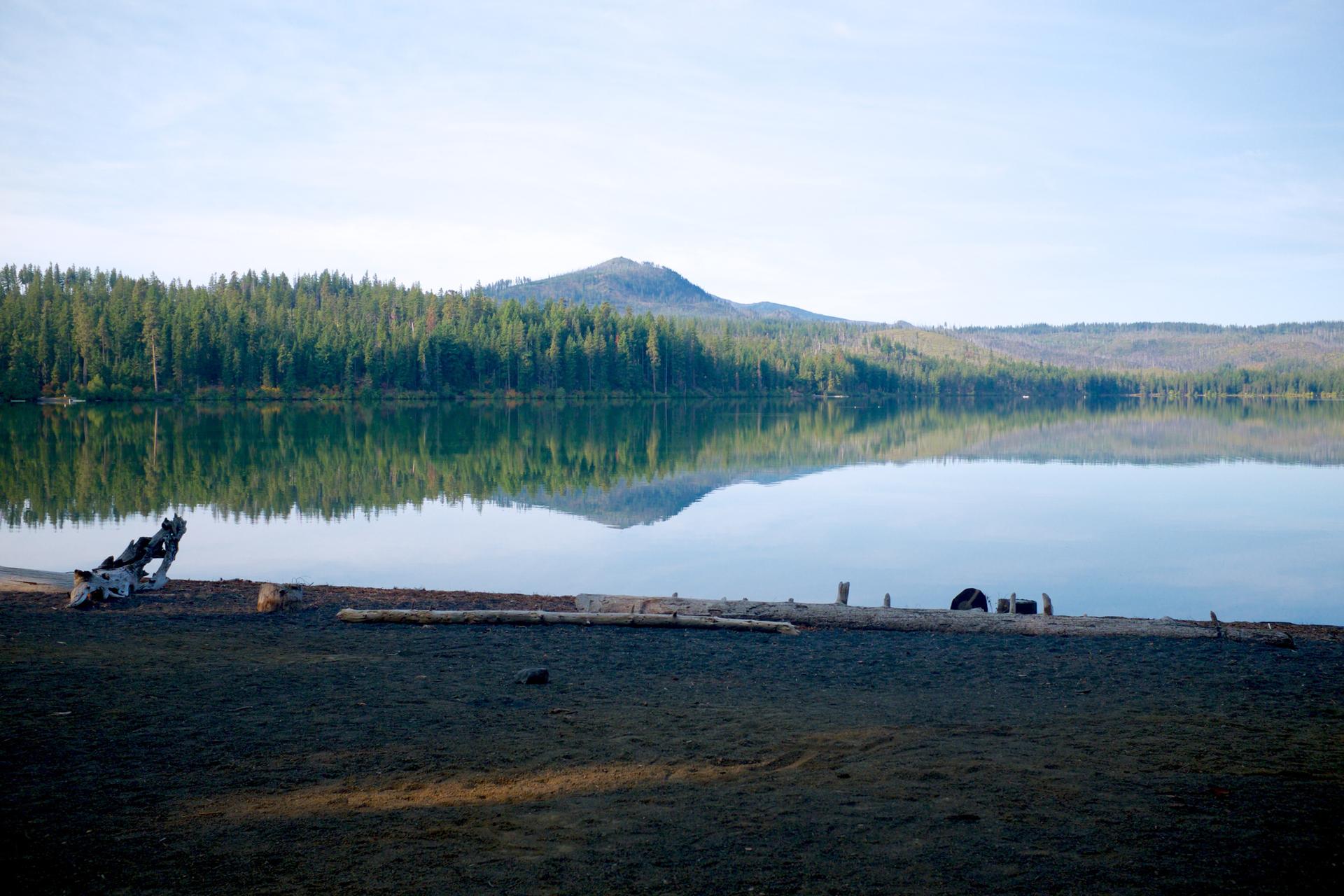 Calm lake reflects forested mountains under a bright sky, with a person on the shore.