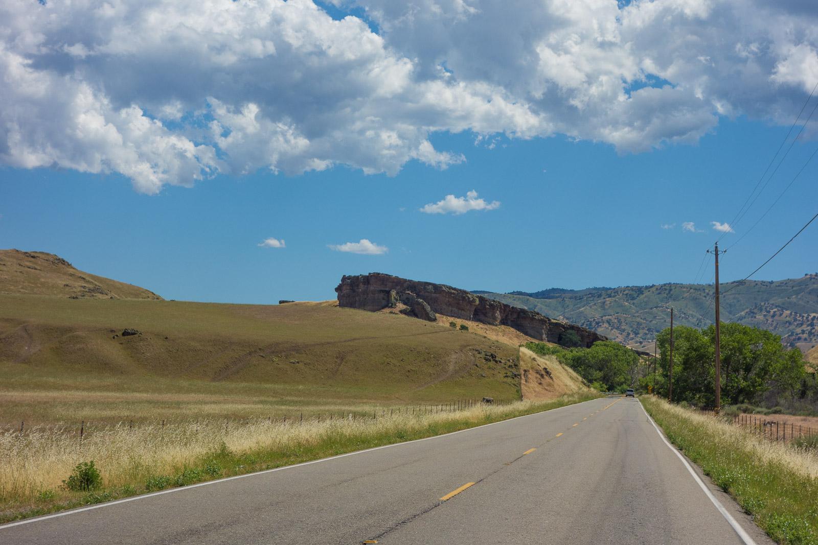 Paved road through grassy fields, hills, and a large rock formation.