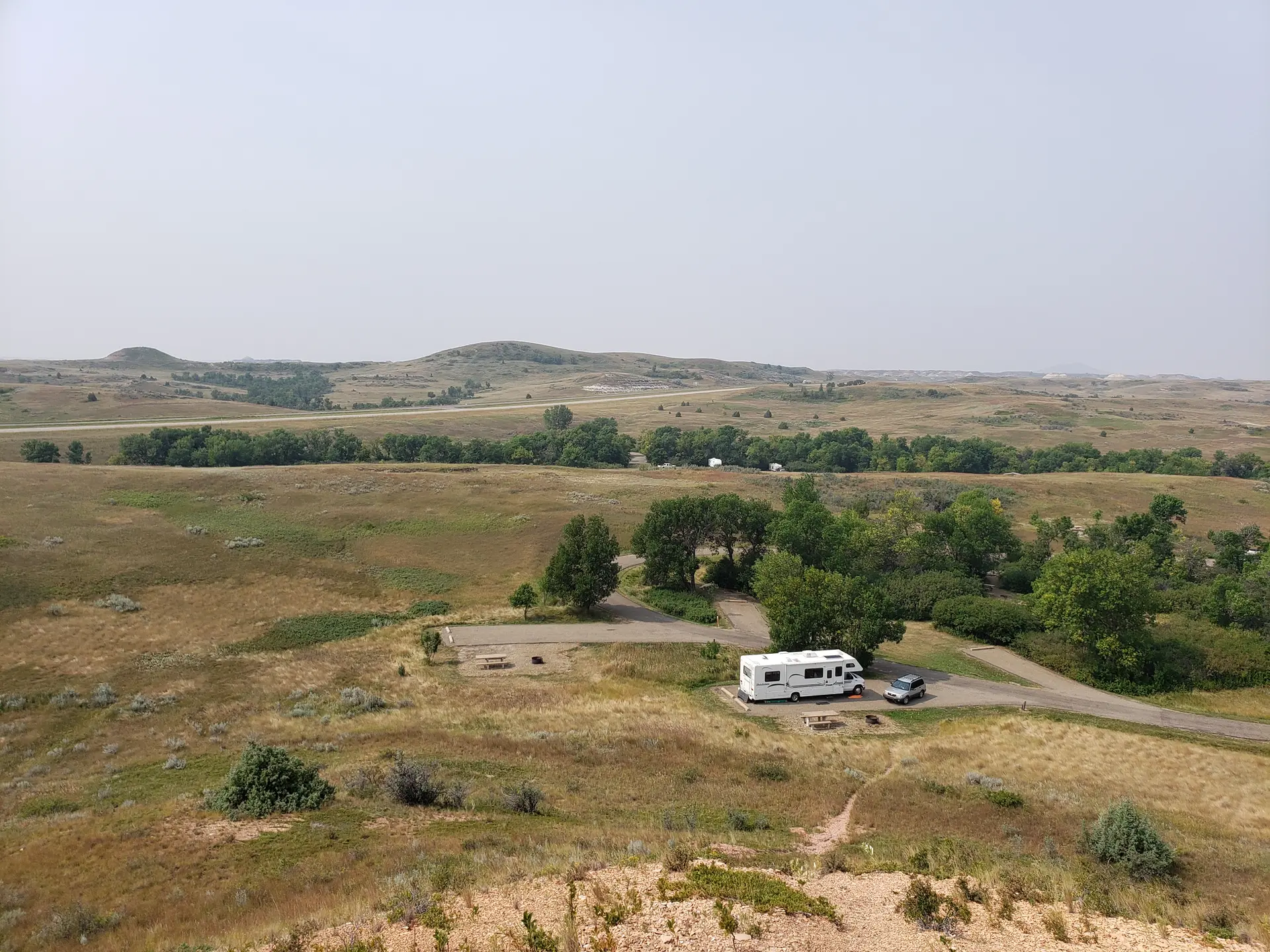 An RV and car parked in a grassy, hilly landscape under hazy daylight.