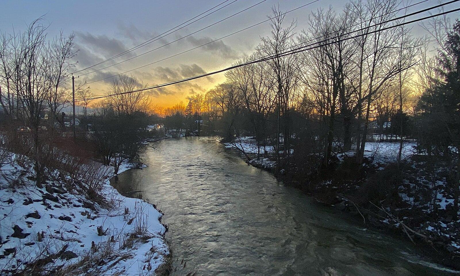 A river flows through a snowy landscape at sunrise with bare trees.