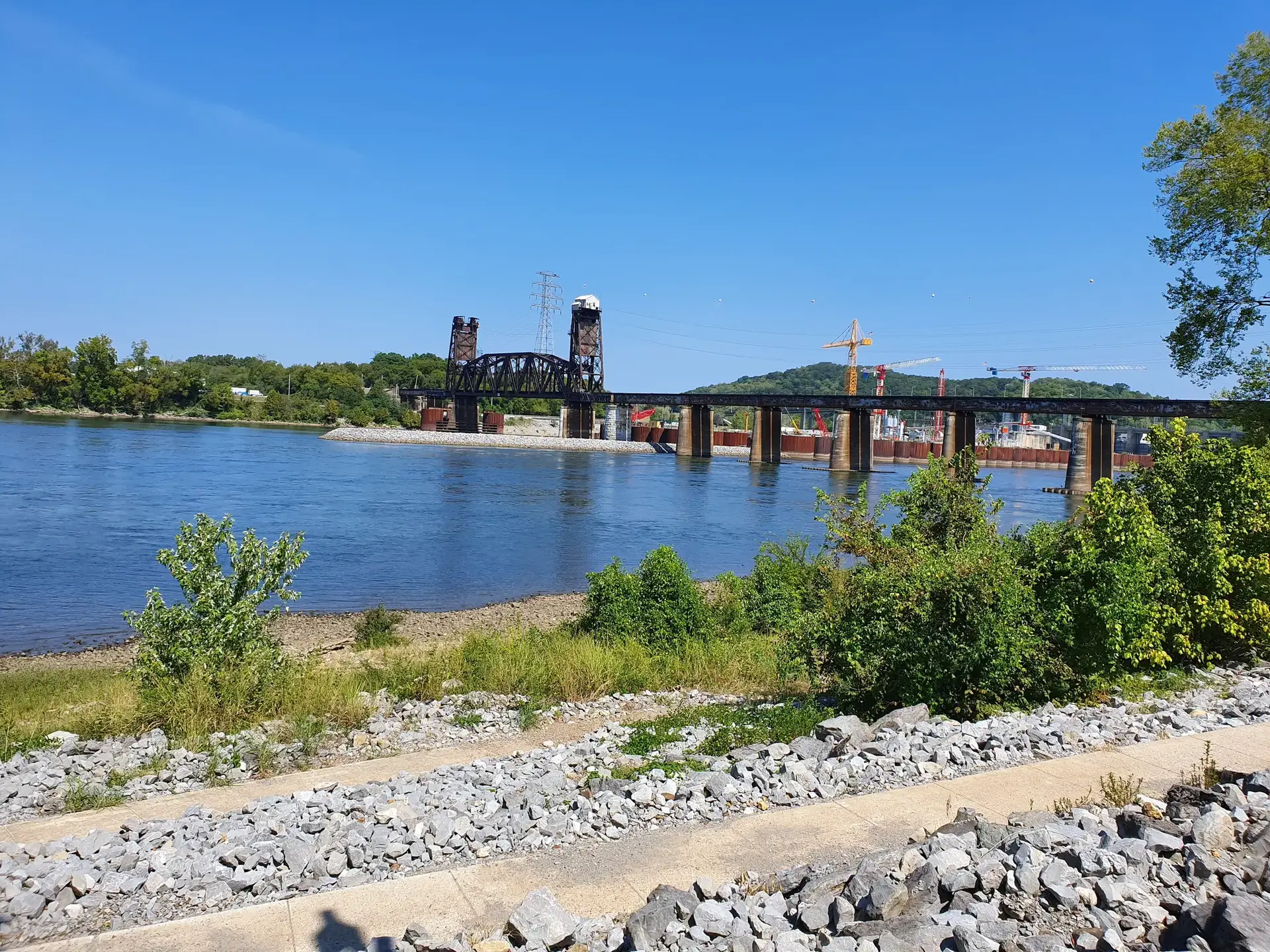 A wide river with two bridges, green hills, and construction cranes under bright sun.