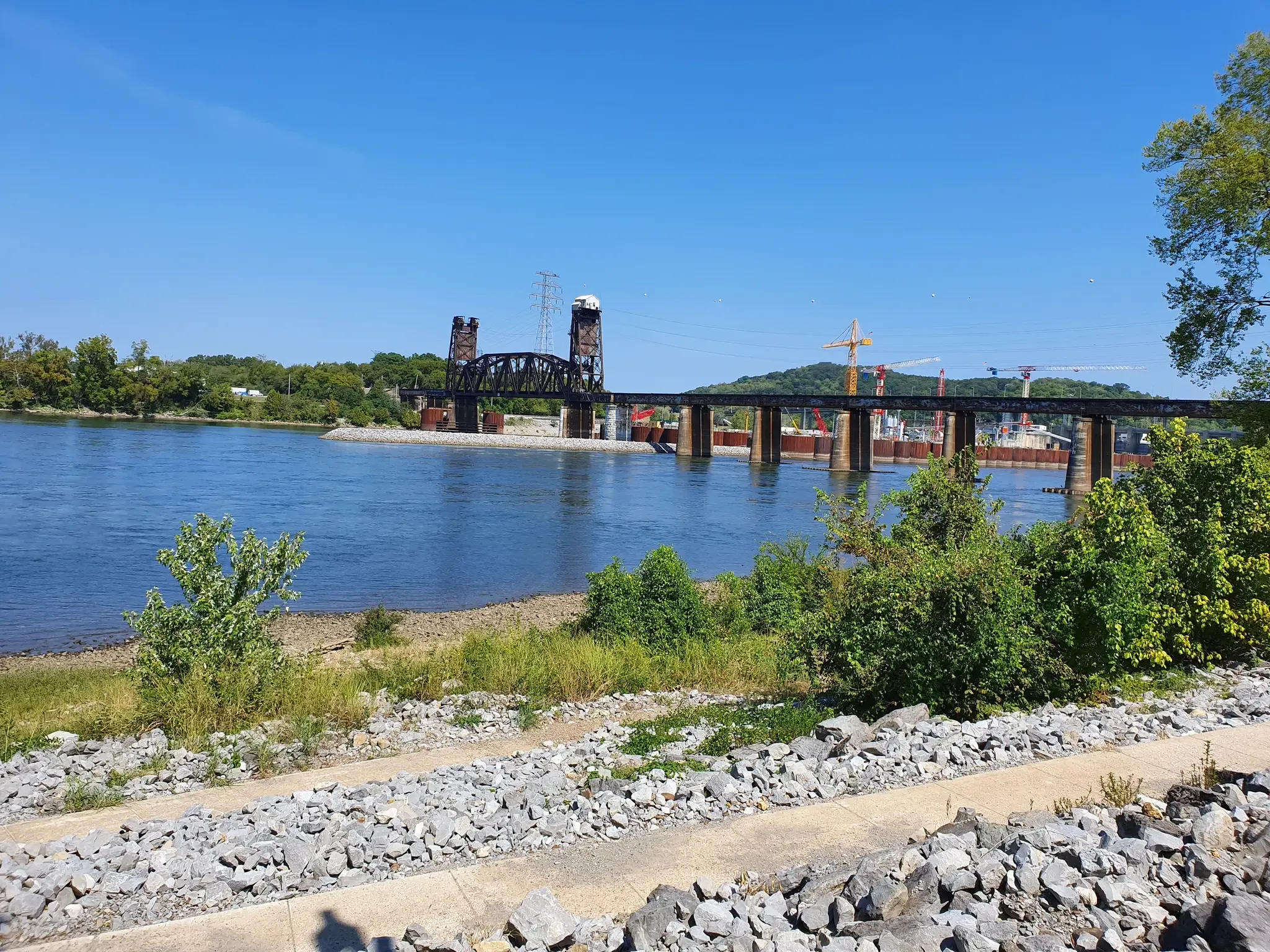 A wide river with two bridges, green hills, and construction cranes under bright sun. - Tennessee Riverwalk Loop