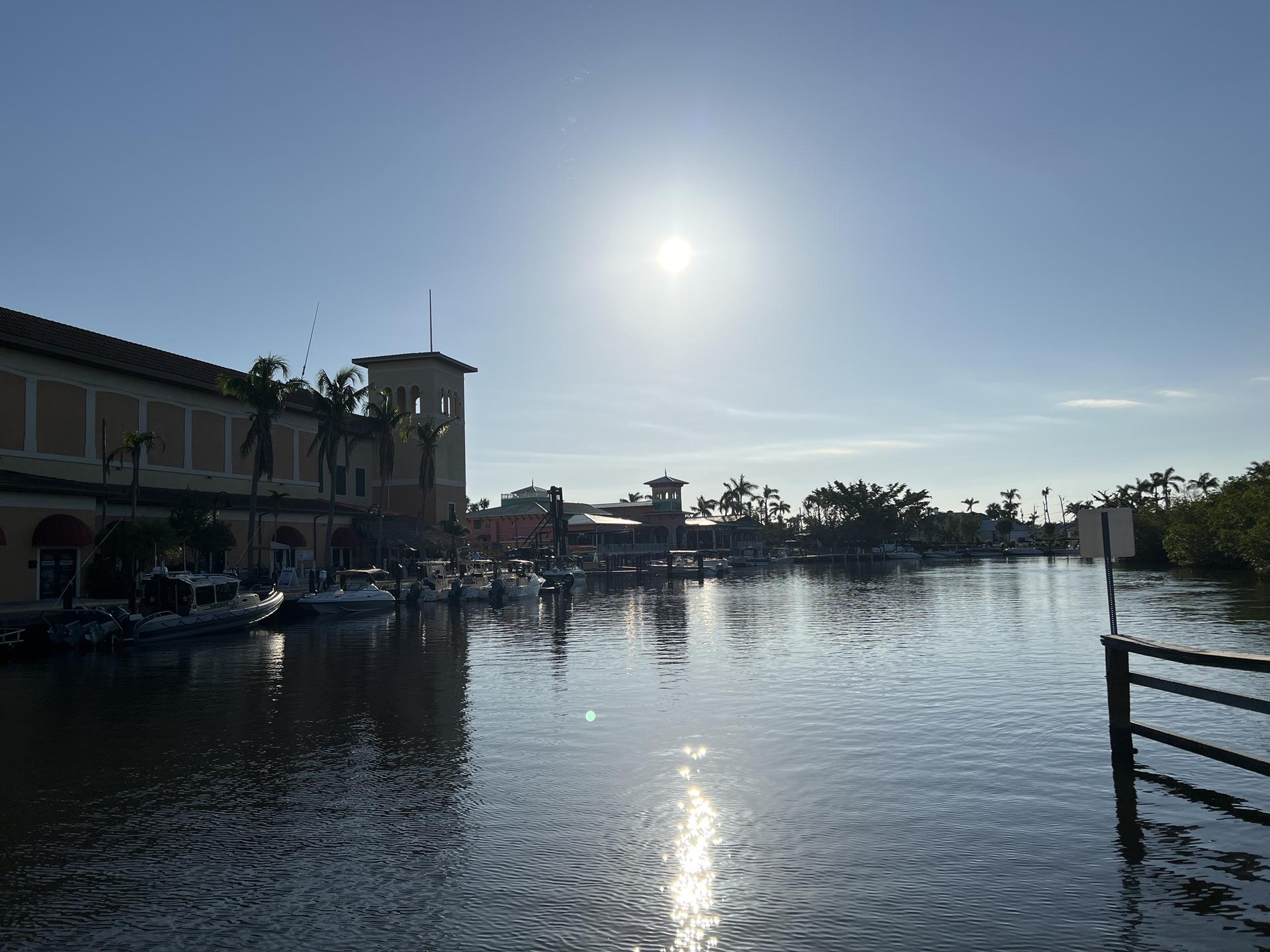 Sun glares over a calm marina with buildings and boats on a bright day.