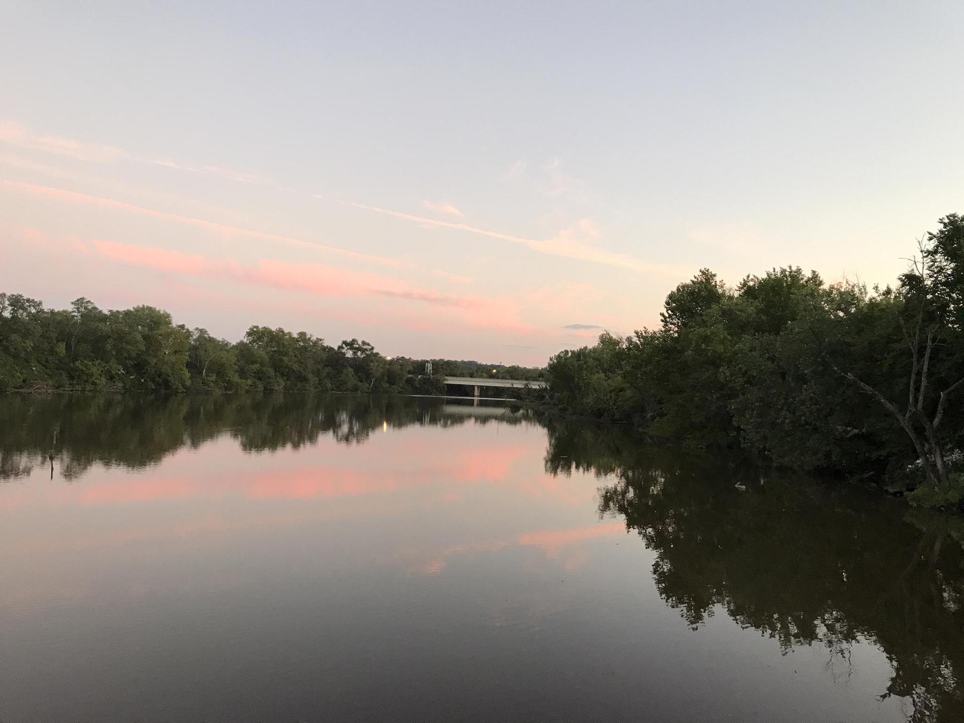 Calm river reflecting pink sunset sky, with distant bridge.
