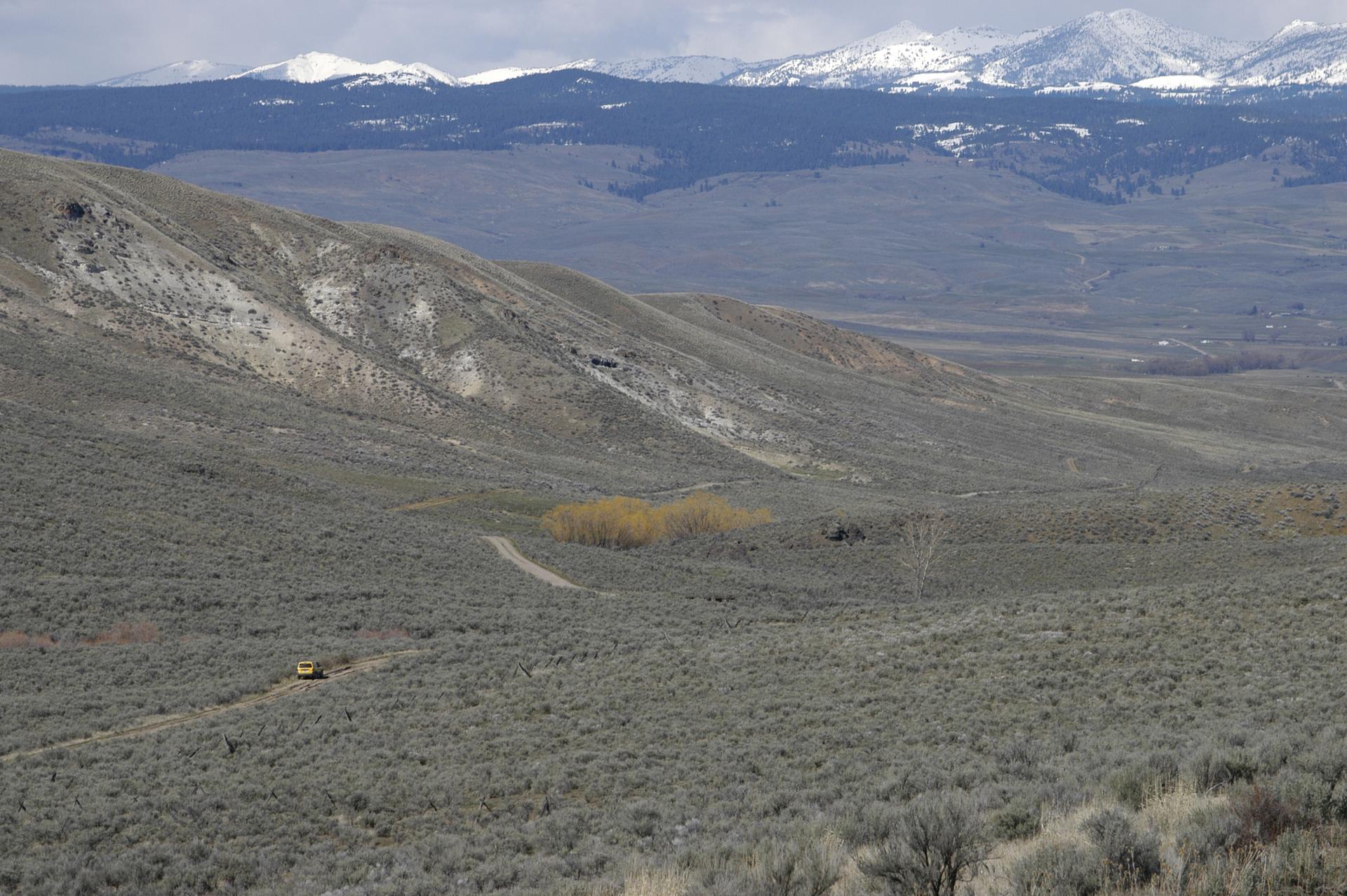 Hilly, arid landscape with a small vehicle and distant snow-capped mountains.