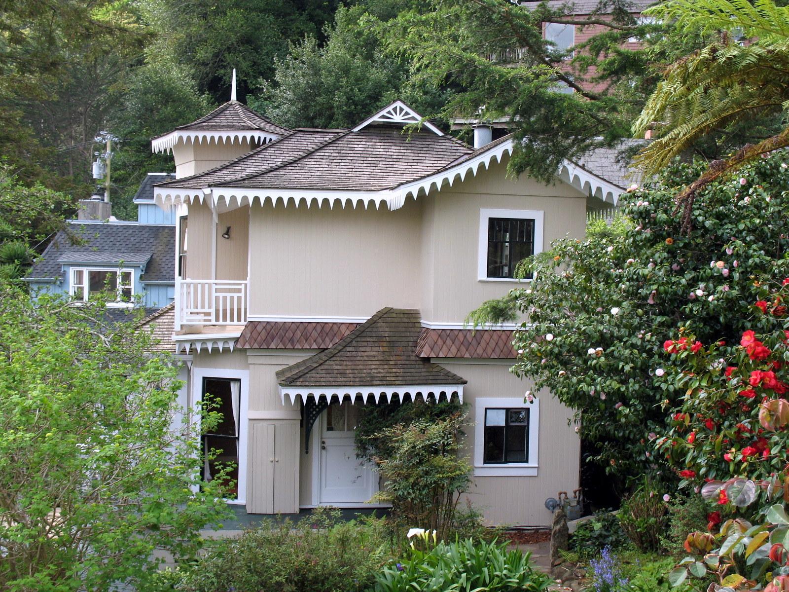 An ornate beige house with decorative trim, surrounded by lush green foliage.