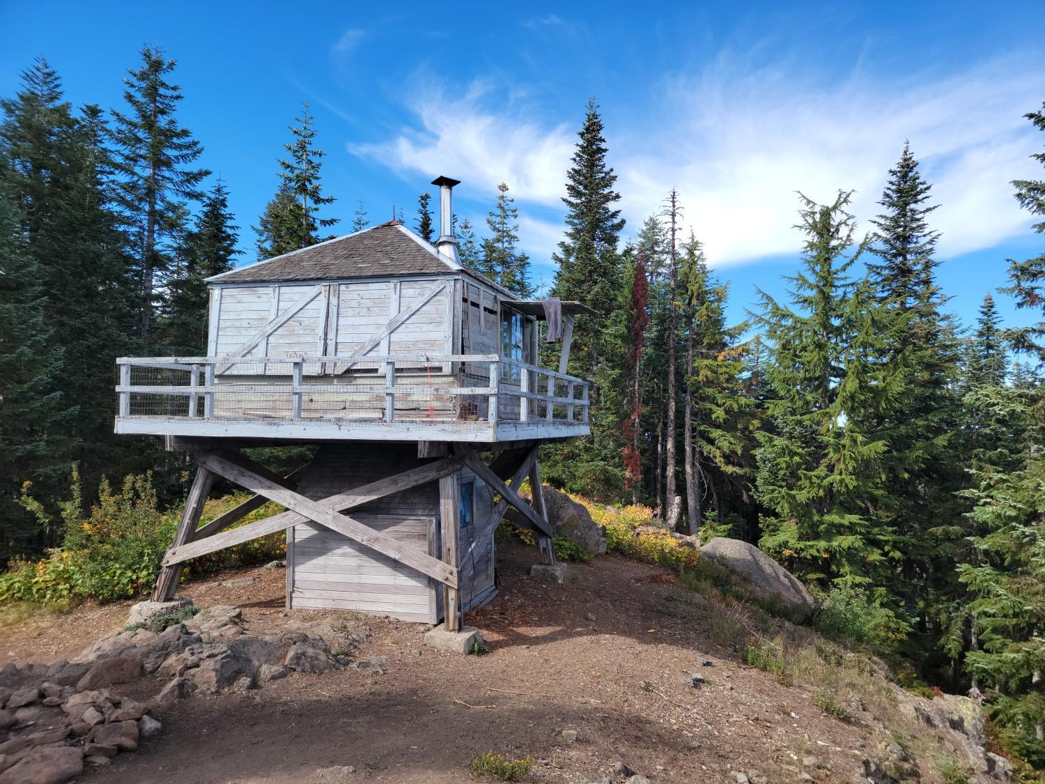 A rustic wooden lookout building stands on a rocky hill among evergreen trees.