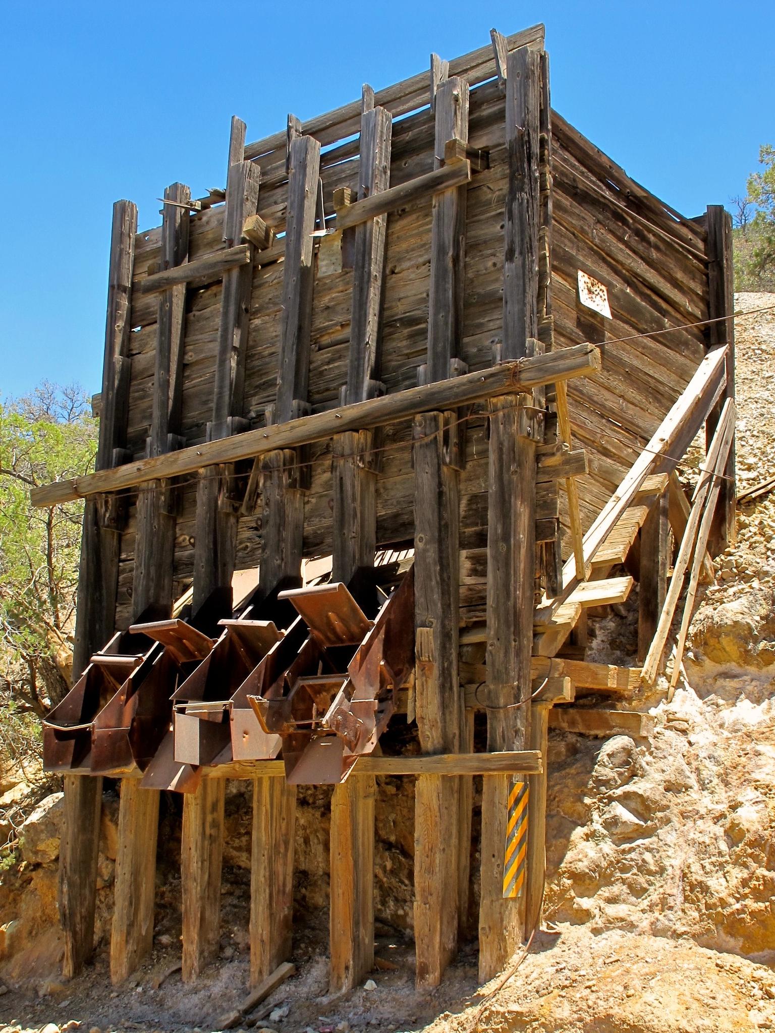 Old wooden mining structure with metal chutes under a bright sky.