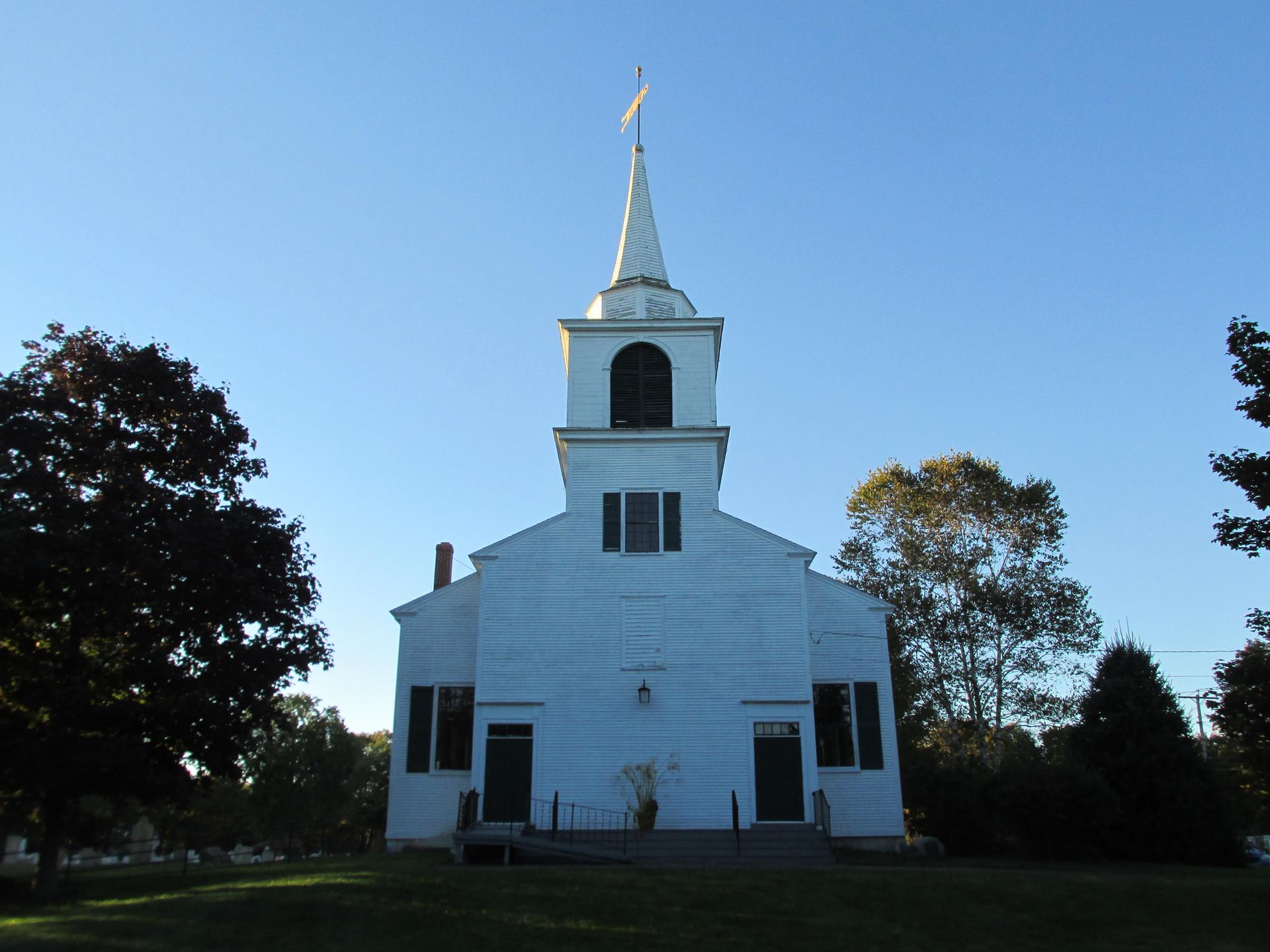 White church with a tall steeple, surrounded by trees, under bright blue sky.