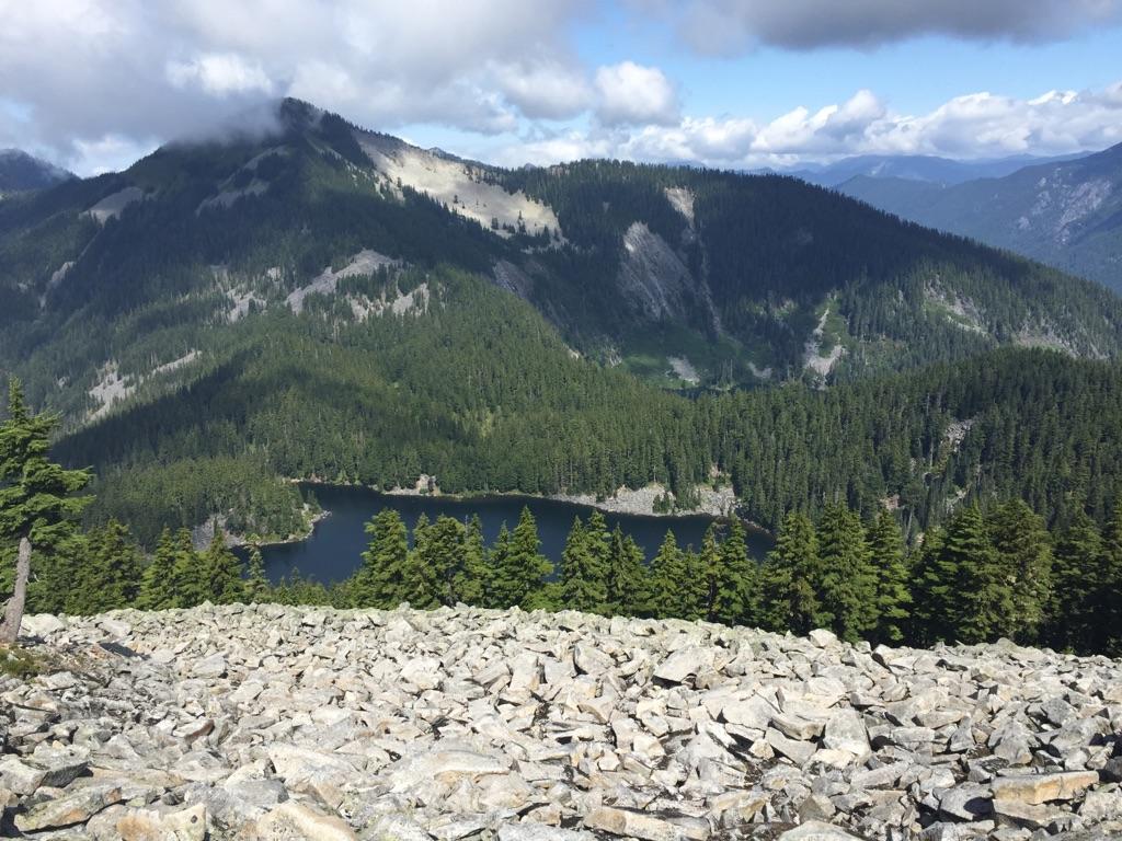 Mountain lake nestled in a forest valley, seen from a rocky, cloudy peak.