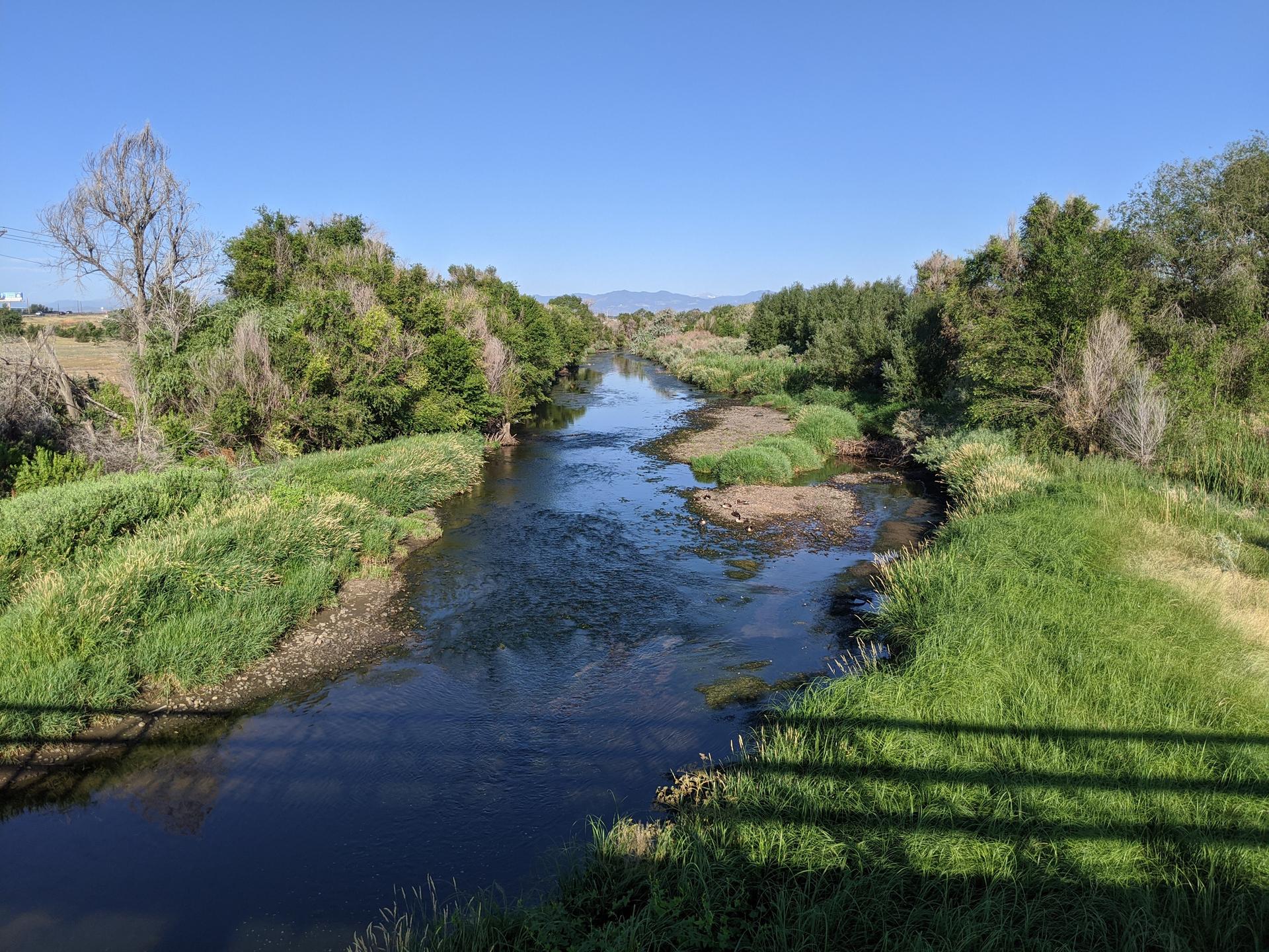 A clear river flows between lush green banks under a bright blue sky.