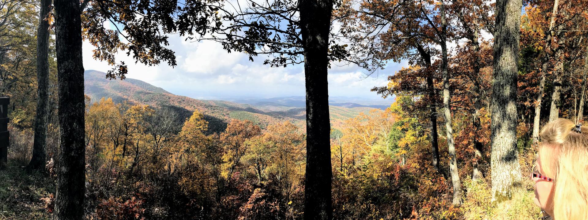Person views vast autumn mountain landscape with colorful trees under a bright sky.