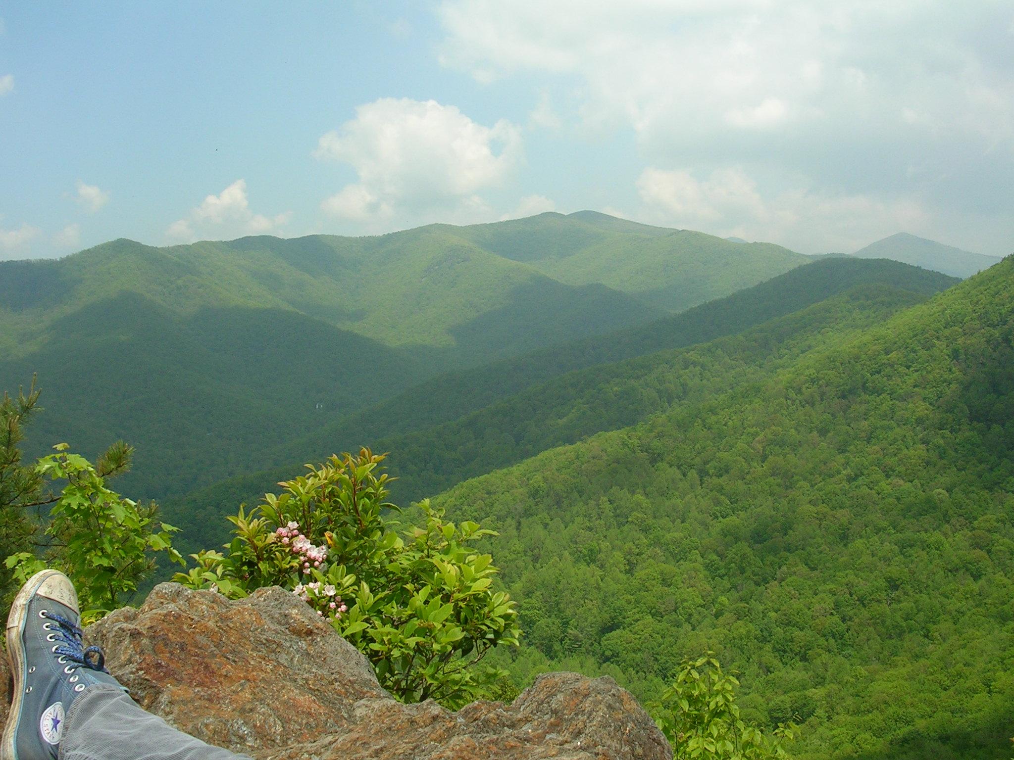 Foot in blue shoe on rock, overlooking green mountains under bright sky.