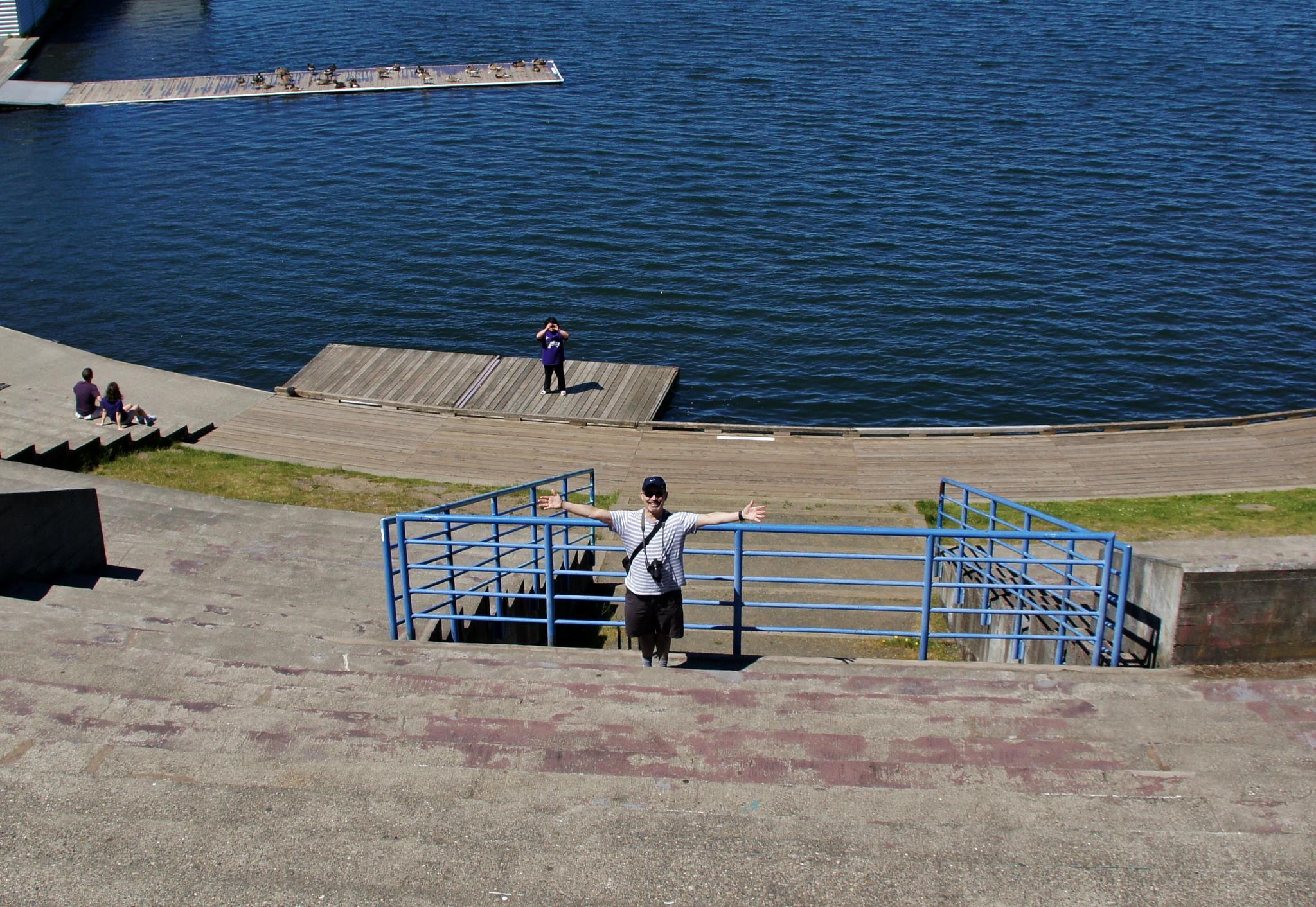 Person posing on concrete steps with a blue lake and wooden dock behind.