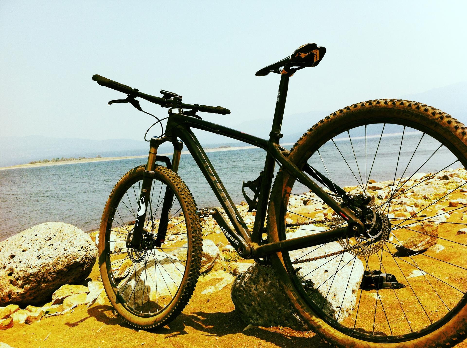 A mountain bike rests on a rocky shore beside calm, sunny water.