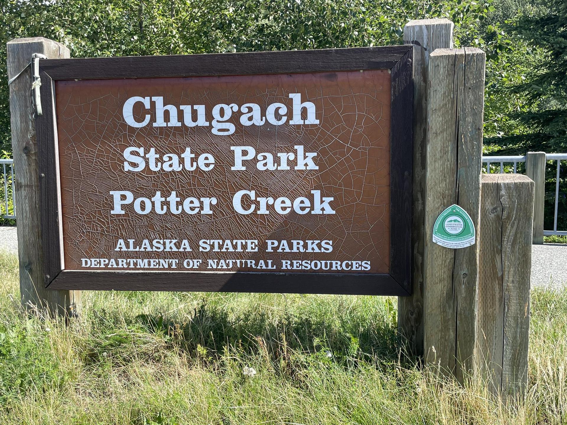 A brown wooden sign with white lettering stands in a grassy outdoor setting.
