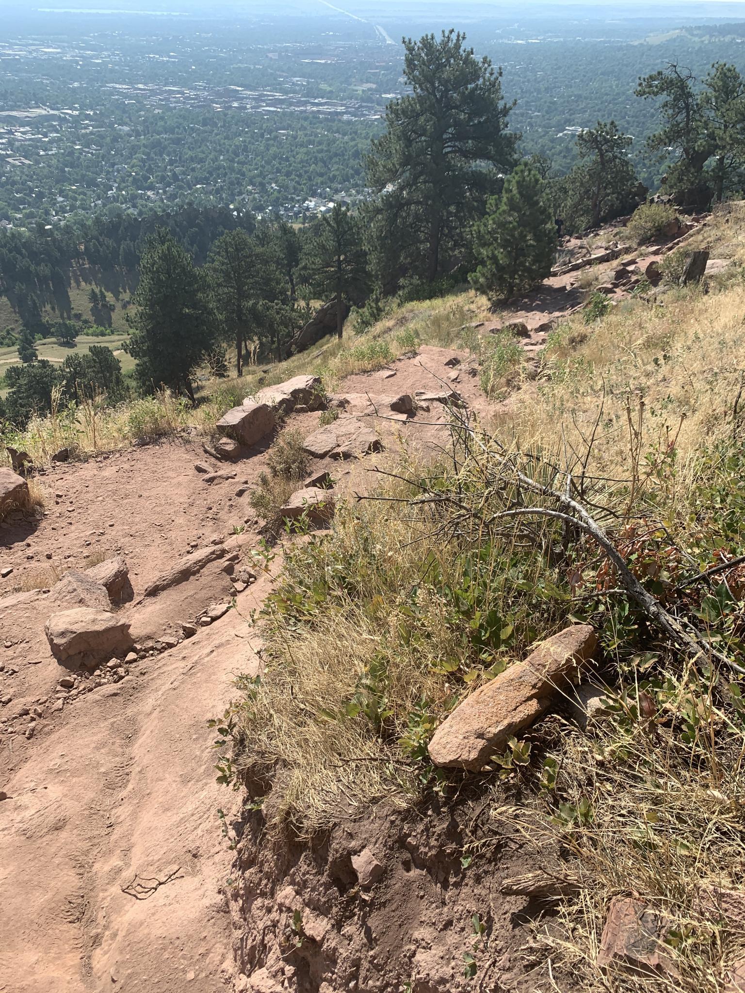A rocky dirt trail winds down a sunny hillside overlooking a distant town.
