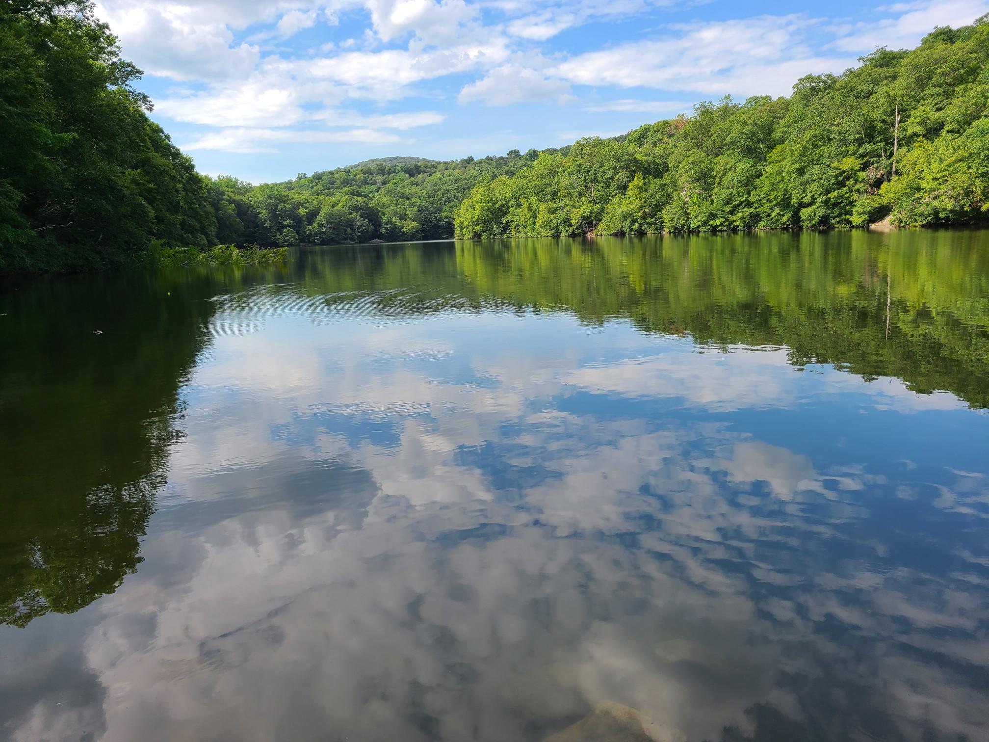 Calm lake reflecting a blue sky with clouds, surrounded by green hills.