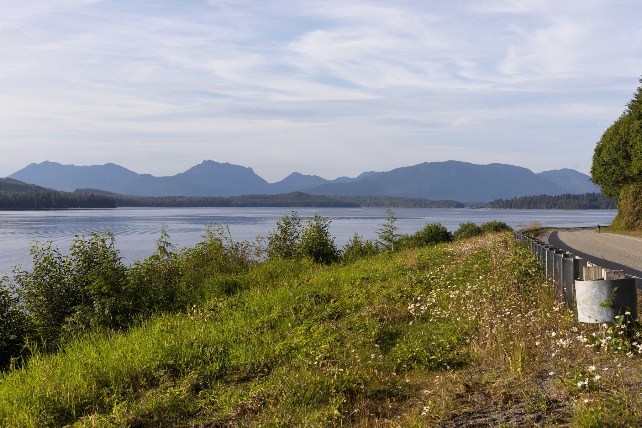 A winding road overlooks a large lake with distant mountains under a bright sky.