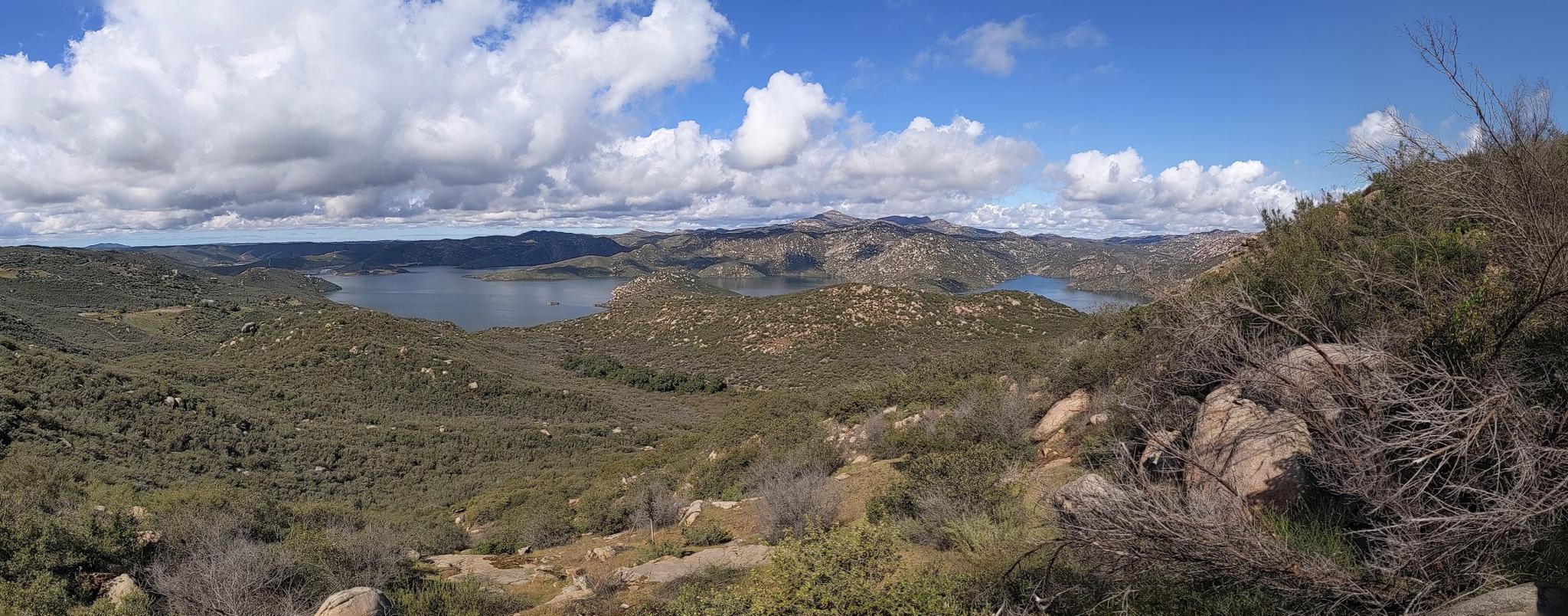Panoramic view of a lake and rocky hills under a bright, partly cloudy sky.